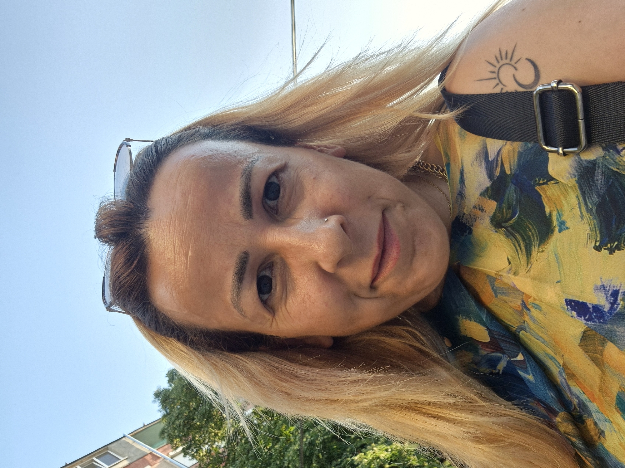 A medium shot captures a woman from the shoulders up, posing for a selfie outdoors against a clear sky. She has long, blonde hair, some of which is pulled back into a bun on top of her head. She is wearing a colorful, abstract-print top with yellow and blue hues, and a black strap across her shoulder. A chain necklace is visible around her neck. A tattoo of a crescent moon and sun is on her left shoulder. Sunglasses are perched on top of her head. In the background, a building with windows and some green foliage are visible.