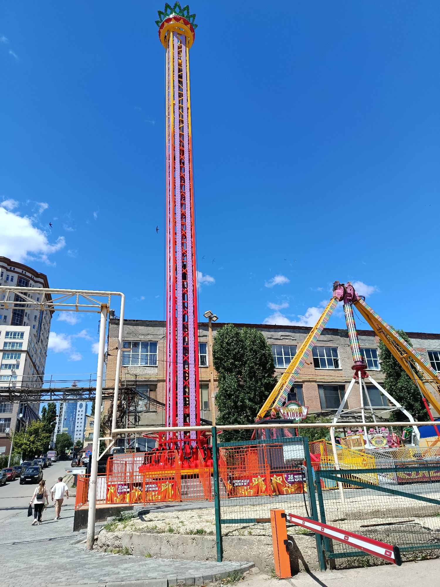 The image depicts an outdoor amusement park on a clear, sunny day. The primary subjects are two large amusement rides: a tall, slender drop tower with a colorful, segmented exterior and a pendulum ride with two large arms that swing in an arc. In the foreground, a red and white barrier gate is lowered.
Two people, a man and a woman, are walking away from the camera on a paved sidewalk. They appear casually dressed for a warm day. Behind them, several cars are parked or driving on a street leading uphill. In the background, there are multi-story buildings, including a modern residential building on the left and an older brick industrial building behind the rides. Trees are visible, adding to the outdoor park atmosphere.
The scene suggests a typical day at an amusement park, with the rides ready for visitors. The bright sunlight and clear blue sky indicate pleasant weather, likely midday or early afternoon. The presence of multiple birds flying in the sky adds a dynamic element to the otherwise static scene.
There is visible text on signage within the amusement park. One sign, partially obscured by the fence, appears to have Cyrillic lettering and the number "36". Another sign with Cyrillic text and graphics is on the front of the fenced-off area. The overall impression is a vibrant and active recreational space under a clear sky.
