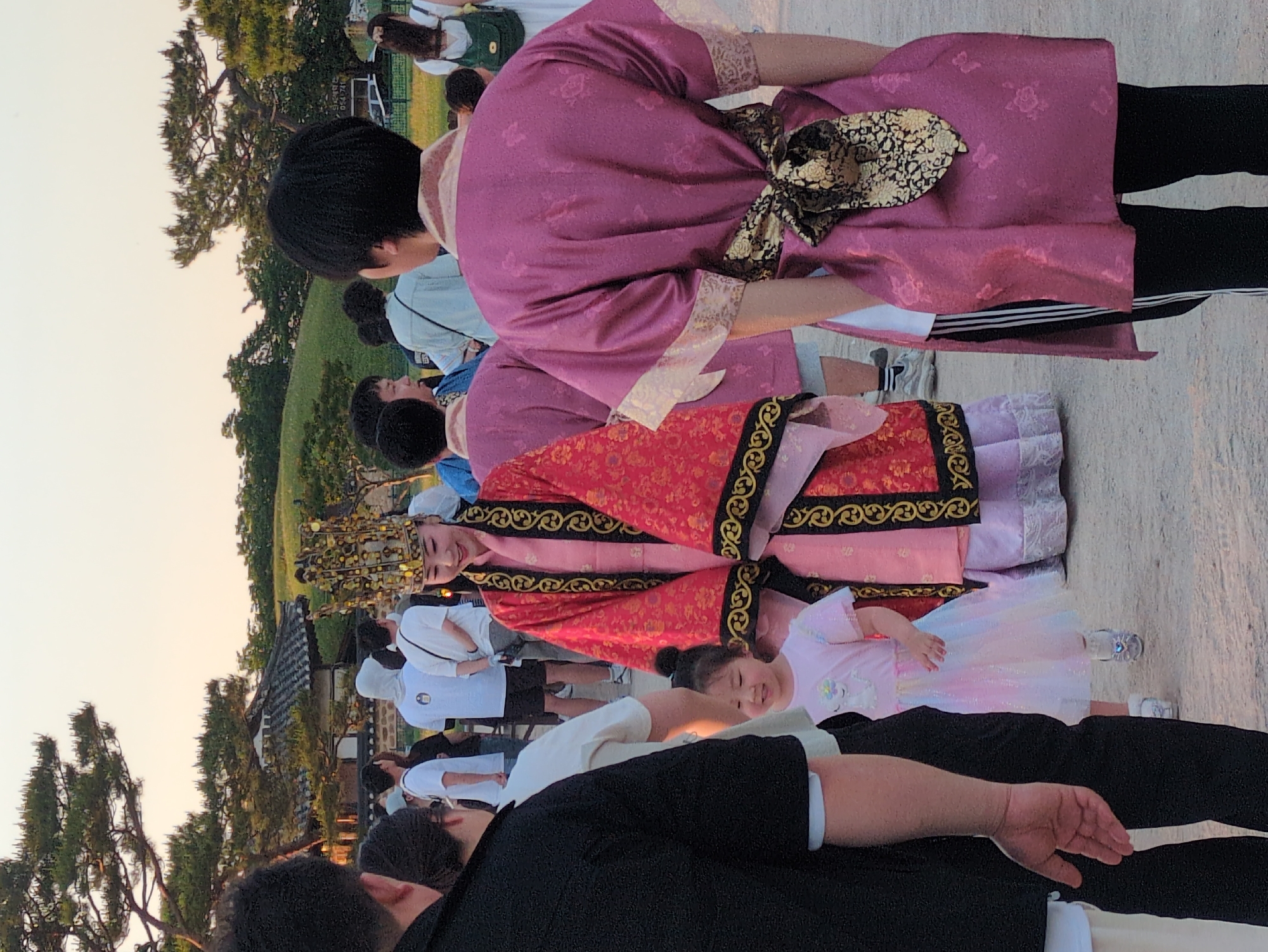 A photograph captures a group of people in Gyeongju-si, South Korea. The foreground contains a person wearing black and white clothing. The background reveals individuals adorned in traditional Korean attire, including a person wearing a red robe with gold detailing and an elaborate headpiece. A young child in a white dress is also visible. The group is assembled outdoors near a traditional Korean-style building. Trees and a grassy hill are included in the backdrop. The sky is a light color, suggesting either early morning or late afternoon.