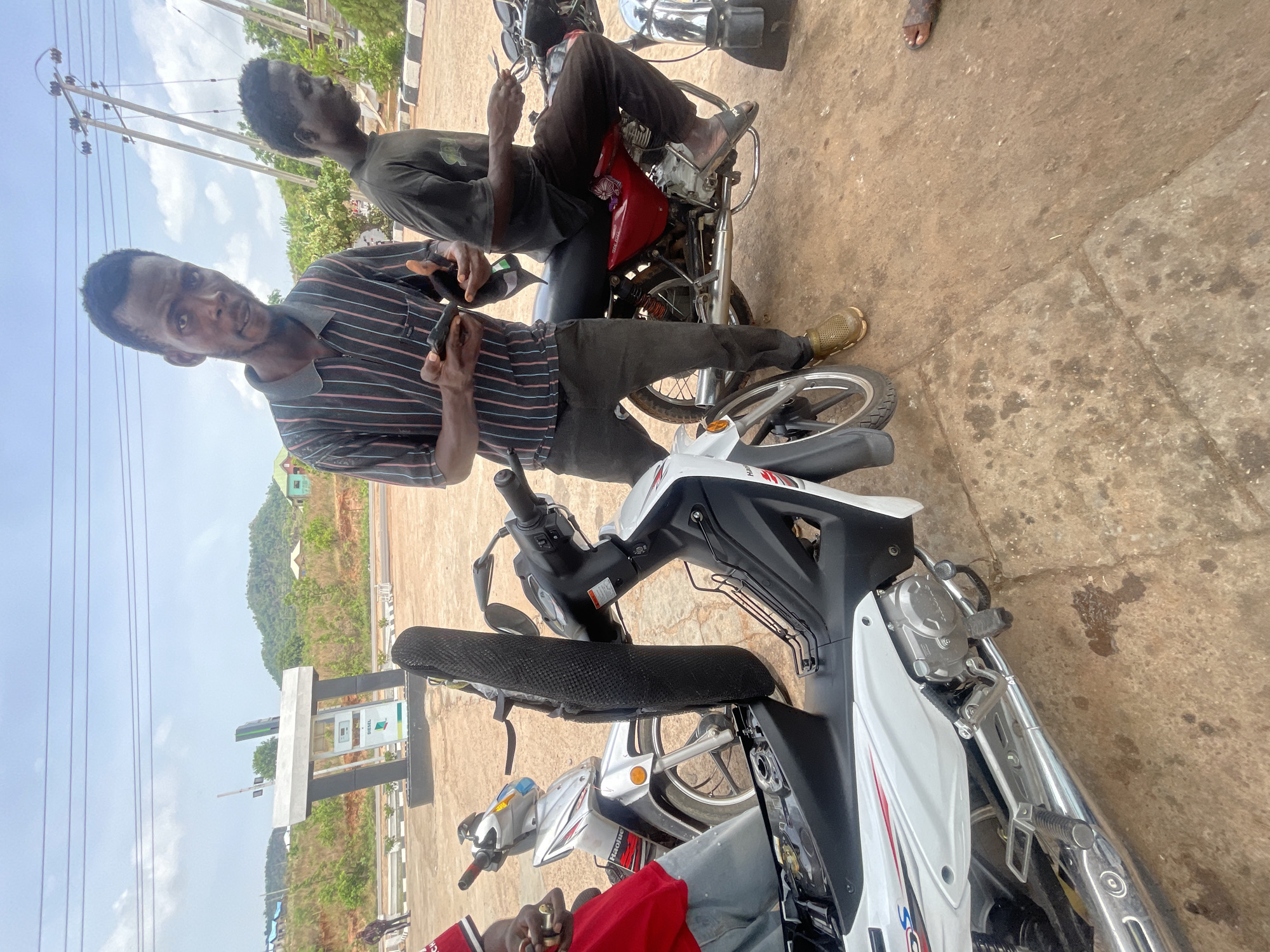 The image shows two men in Lokoja, Nigeria, standing next to motorcycles. One man, wearing a striped shirt and dark pants, holds a small object in his hands. The other man, wearing a dark shirt, sits on a red motorcycle and looks at his phone. In the background is a gas station and green hills.
