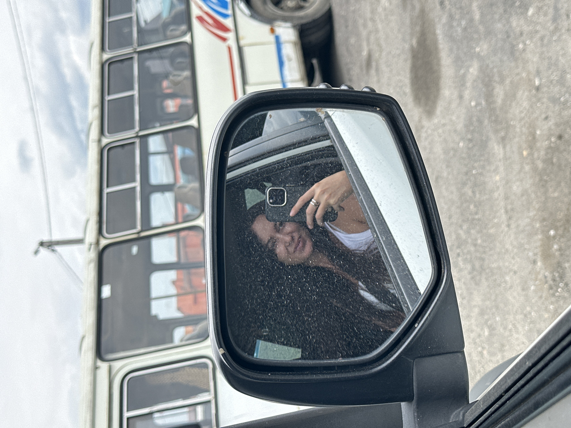 The image shows a woman taking a selfie in a car's side mirror. She is wearing a white tank top and has long, dark hair. She is holding a smartphone in her right hand and wearing rings on her fingers. In the reflection of the mirror, a white bus with red and blue stripes is visible in the background. The environment appears to be an outdoor setting, possibly a street or a parking lot, with a cloudy sky overhead. The specific location in the city of Slyudyanka, Russia, is not identifiable from the visual information provided in the image.