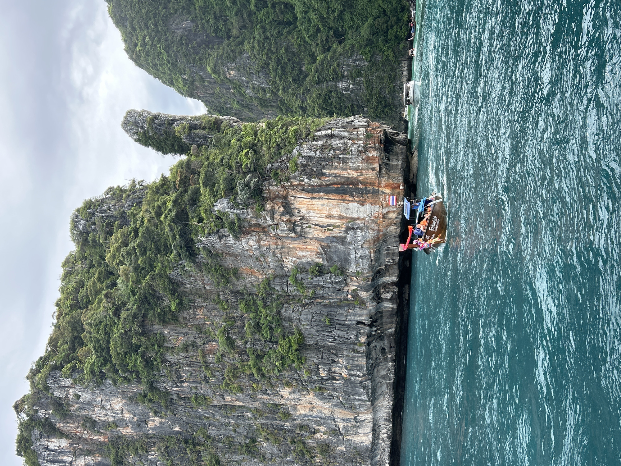 This is a photograph taken from a boat on turquoise water, looking towards a massive, jungle-covered limestone cliff face. A traditional longtail boat, carrying several people, is in the foreground, moving away from the camera. Several other boats are visible in the distance, some white motorboats and more longtail boats. The sky is overcast with clouds. The scene depicts a view of the karst landscape characteristic of Ao Nang, Thailand, likely in the Phi Phi Islands or a similar coastal area accessible from Ao Nang.