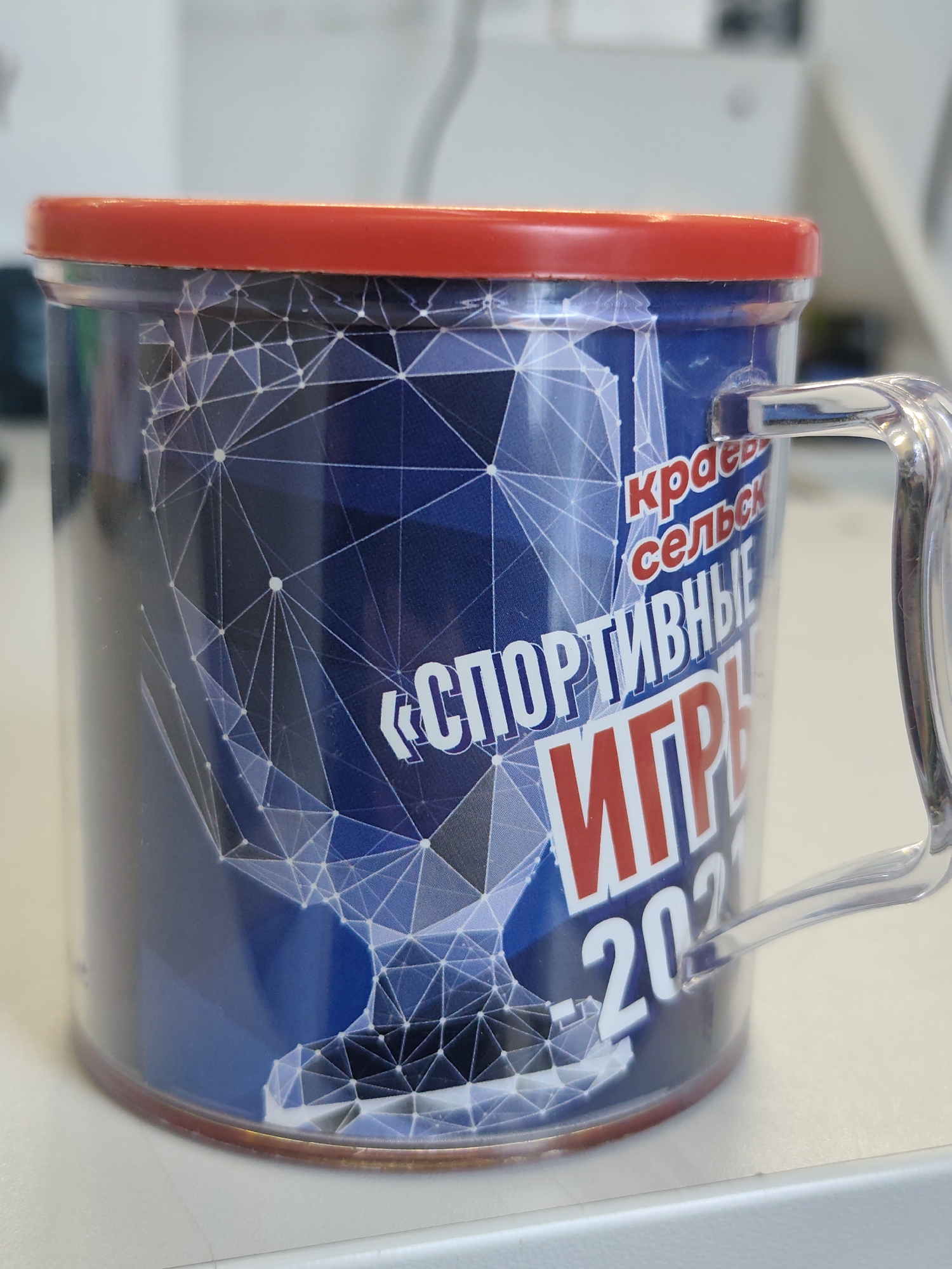 A cylindrical mug with a red lid and a transparent handle is placed on a white surface, likely a desk. The mug is predominantly blue with a white geometric pattern resembling a network or constellation. Text in white and red is visible on the mug's surface, including what appears to be Russian text. The red text reads "КРАЕВЫЕ" (KRAEVYE) and "СЕЛЬСК" (SEL'SK), and the white text reads "«СПОРТИВНЫЕ»" («SPORTIVNYE») above a large red word that appears to be "ИГРЫ" (IGRY), meaning "GAMES." Below the word "GAMES" is the year "202" followed by another digit. The mug is positioned at an angle, with the handle extending to the right. The background is blurred but suggests an indoor environment with white and light-colored elements, possibly office furniture or walls. There are no people or animals visible in the image. The scene does not provide any specific information to indicate its location within the city of Chaikovsky, Russia.