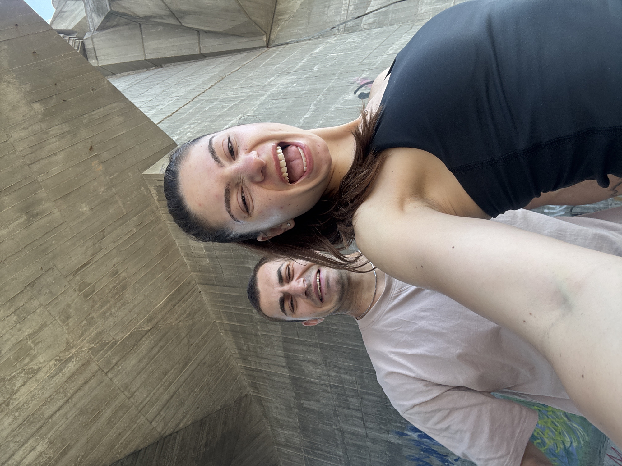 Two individuals, a man and a woman, are pictured from the chest up, taking a selfie. The woman, in the foreground, is making a comical face, with her mouth wide open and her eyes squinting. She is wearing a black tank top. The man, in the background, is also making a distorted facial expression, with his eyes squeezed shut and his mouth pulled into a grimace. He is wearing a light pink t-shirt and a silver chain. They are standing under a large concrete structure, possibly an overpass or a bridge, with visible graffiti on the wall behind them. The lighting suggests it is daytime. This scene is likely located in Varna, Bulgaria, under the bridge of the pedestrian passage leading to the city center from the northern districts, often referred to as the "concrete jungle" due to the surrounding architecture.