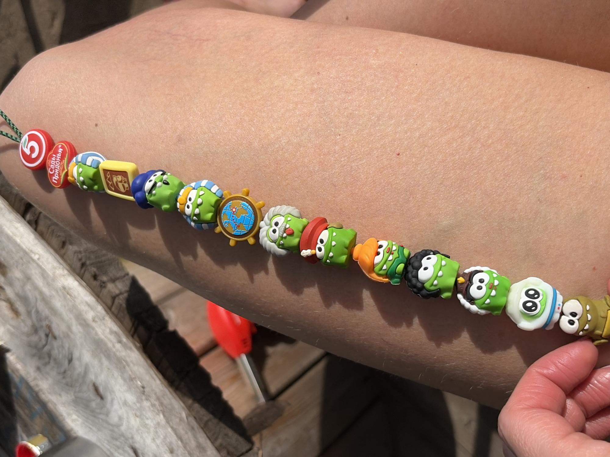 Here is a detailed description of the image:
**Scene:** The image shows a close-up view of a bracelet made of various colorful charms. The bracelet is resting on a person's forearm or upper arm. The background includes a wooden surface, possibly part of a deck or outdoor furniture.
**Subjects/Objects:**
* **Bracelet:** The primary focus is the bracelet, which appears to be made of rubber or plastic charms strung together on a thin green cord.
* **Charms:** The charms have various designs, including cartoon-like figures, possibly monsters or other characters, as well as other shapes like a ship steering wheel with a globe in the middle. There is also a red plastic piece and another red piece that say "Сады Придонья" in Russian, and a yellow square piece.
* **Arm:** A person's arm is visible, showing skin with some visible pores and minor blemishes.
* **Hand:** A portion of a hand is visible in the lower-right corner of the image.
* **Wooden Surface:** The background includes weathered wooden planks or boards.
* **Red Object:** A red object is visible between the arm and the wooden surface.
**Actions/Interactions:**
* The bracelet is placed on the arm, but there is no specific action occurring in the image.
**Environment:**
* The lighting suggests it is daytime, possibly outdoors.
**Location:** The presence of the Russian text "Сады Придонья" suggests that this photo may be taken in Russia. There is no definitive evidence to place this picture in the specific city of Забегаево, Russia, as the Russian text would be available to the public. More context would be required to ascertain this location.