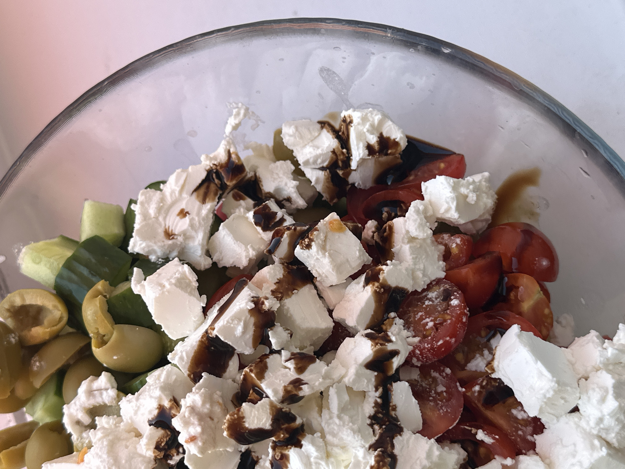 The image shows a close-up of a salad in a clear glass bowl. The salad contains green olives, sliced cucumbers, halved cherry tomatoes, and cubed feta cheese. A dark sauce, possibly balsamic glaze, is drizzled over the cheese. The background is blurred and indistinct. The image was not necessarily taken in the city of Chaikovsky, Russia.