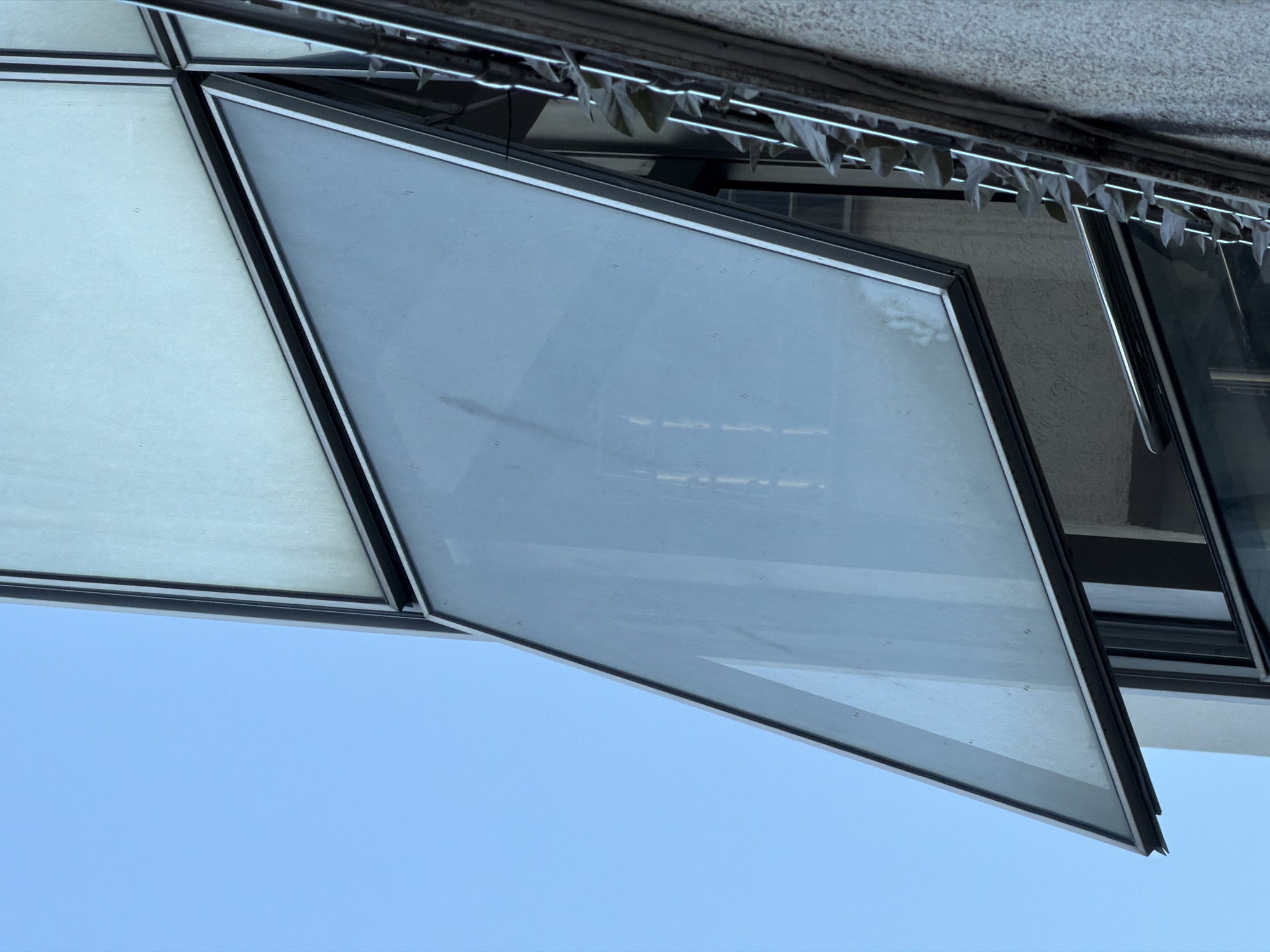 An upward-angled shot reveals a building facade in Budva, Montenegro, showcasing a geometric arrangement of windows set against a clear blue sky. The architectural design features irregularly-shaped windows, some opaque and others transparent, framed in a dark metal. The nearest window is angled outward, suggesting it is in an open position. Above the windows, a textured, light-colored exterior surface is visible, with a string of small, possibly decorative, items hanging along the upper edge. Adjacent windows reflect the sky and surrounding structures, adding to the complexity of the architectural composition. No people are visible in the frame.