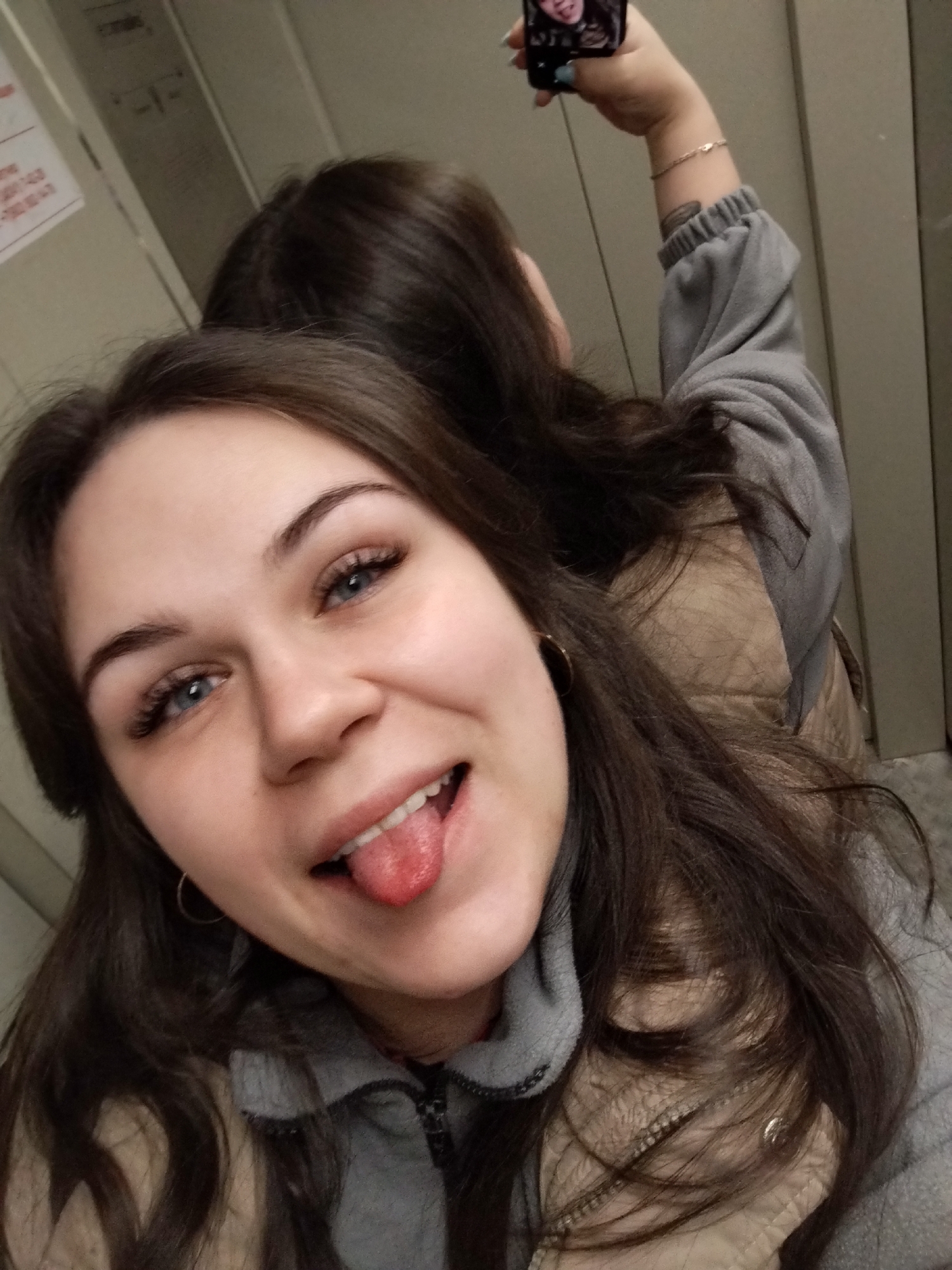 Two individuals are captured in a close-up selfie within an indoor, confined space, possibly an elevator, in the city of Chaikovsky, Russia. The walls are light-colored and paneled, with a partially visible white notice containing red Russian text and numbers (e.g., "07-00", "08-00") affixed in the upper left background.
In the foreground, a young female with long dark brown hair and blue eyes looks directly at the camera, smiling with her mouth open and tongue extended in a playful manner. She wears a grey zipped fleece jacket layered over a beige puffer-style vest and has small gold hoop earrings.
Behind and slightly to the right of the foreground individual, a second female is partially visible. Her right arm is raised, holding a black smartphone overhead to take the photograph. She wears a grey long-sleeved top, a thin bracelet is visible on her wrist, and a dark marking or tattoo is present on her forearm. A reflection of a face, likely the foreground person, is visible on the smartphone screen.