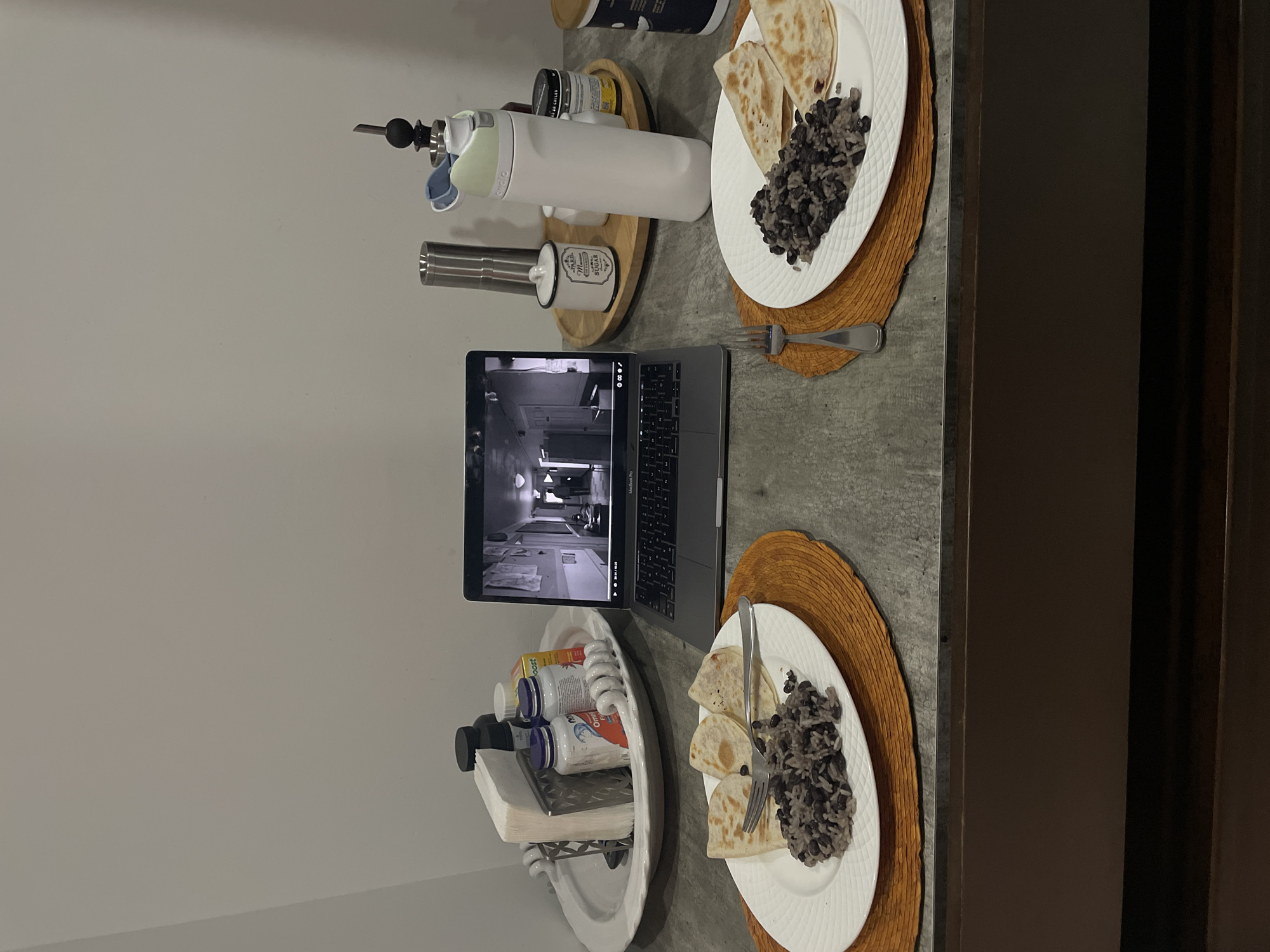 A table setting displays two plates of food, each containing quesadillas and black beans. A laptop is positioned between the plates, displaying a video in black and white. Various items are placed around the laptop and plates, including bottles, containers, and kitchenware. The location of the scene is in Huixquilucan de Degollado, Mexico.