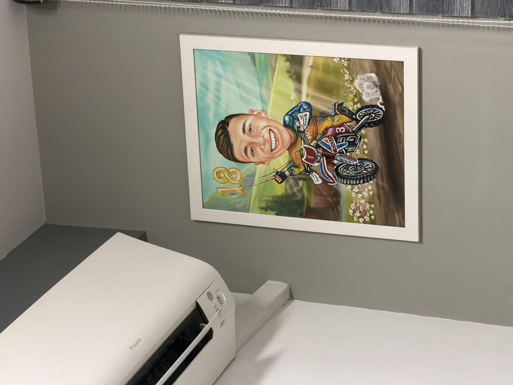 The image captures an indoor scene, primarily featuring a framed caricature mounted on a solid grey wall. The caricature depicts a young man with a large, jovial head, short dark hair, and a wide, toothy smile. He is clad in a colorful motocross racing suit, predominantly blue, yellow, and red, with "ONE" partially visible on his chest and the number "3" displayed on the side of his dirt bike. The man is holding a golden balloon shaped like the number "18," suggesting a birthday celebration. The background of the caricature is a vibrant, sunlit landscape with green fields, rolling hills, and patches of white and pink flowers.
In the foreground, towards the bottom left, a white Daikin air conditioner is visible, resting on a white surface, possibly a shelf or desk. Above the framed picture, along the top edge of the wall, there is a horizontal fixture that appears to be a blind or curtain rail with an integrated LED light strip. The overall lighting suggests a well-lit environment, emphasizing the cheerful expression of the caricature subject.