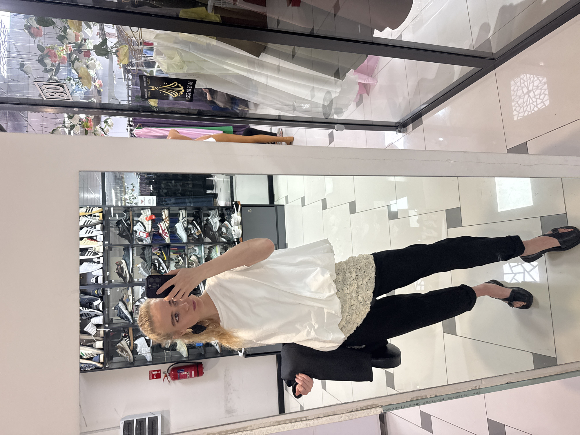 The photograph captures a woman taking a selfie in front of a mirror. She is seated on a black stool and is wearing a white blouse with a detailed trim, black trousers, and black sandals. Her blonde hair is tied back, and she is holding a smartphone to capture the image. The mirror reflects a display of shoes, predominantly sneakers, arranged on shelves. To the left of the display is a red fire extinguisher. The flooring consists of a patterned tile arrangement. A reflection of a retail display, complete with hanging clothes and floral decorations, is visible near the top edge of the frame, implying the store environment.