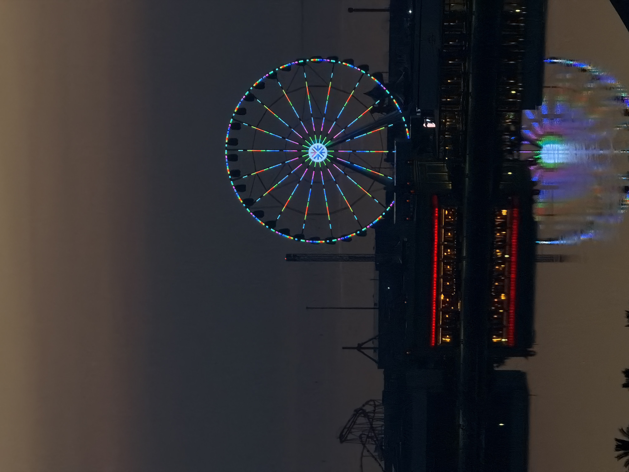 The image captures a Ferris wheel illuminated with colorful lights against a dusky sky. The Ferris wheel is the central focus, with its spokes and cabins clearly visible. Below the Ferris wheel, a building with red lights is illuminated, and its reflection is visible in the water in the foreground. There are also structures resembling roller coasters in the background on the left. The overall scene suggests a nighttime view of an amusement park or entertainment venue.