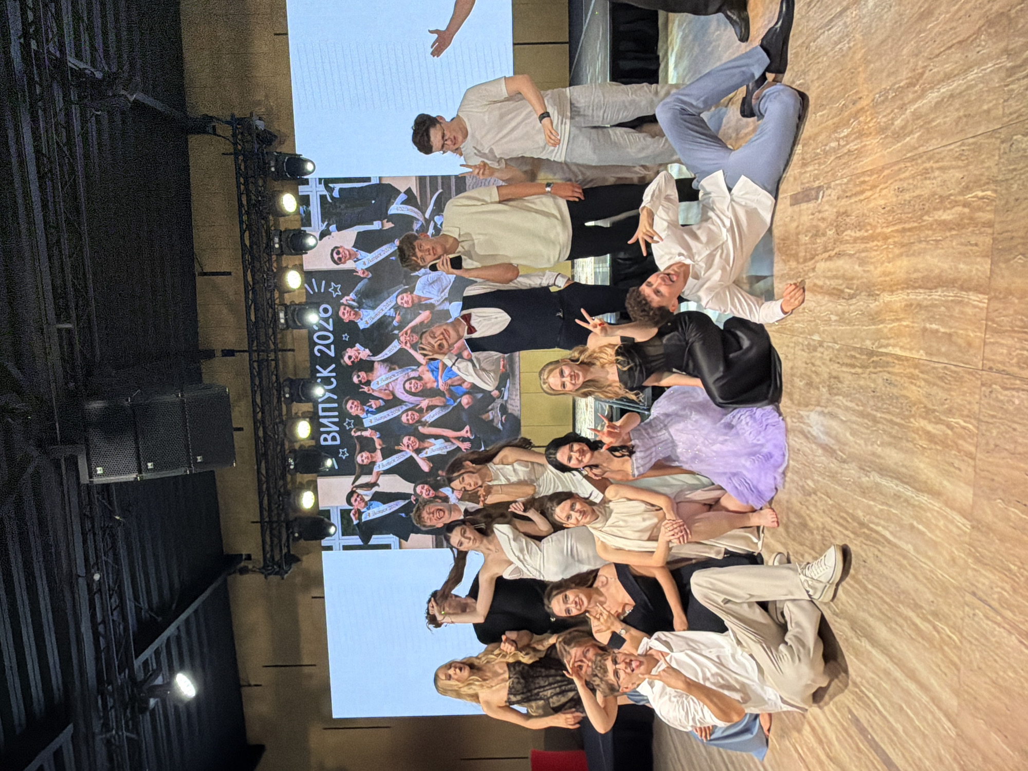 This media file shows a large group of young adults, likely students, posing for a photo on a stage. They are dressed in formal attire, with many men wearing white shirts and suits, and women in dresses. Some are wearing sashes that appear to say "Випуск 2020" (Graduation 2020). The background features a large screen displaying images of other students, also likely from the same graduation event. Lighting equipment, including spotlights, is visible on trusses above the stage. The floor is a light-colored marble or stone pattern. The overall atmosphere appears celebratory, with smiles and gestures of excitement among the individuals. The image is taken indoors, likely at a school or event venue, given the stage and screen setup. The lighting suggests it is likely evening or a dimly lit indoor space. While the exact location is not specified in the image itself, the context provided states Khotianivka, Ukraine.