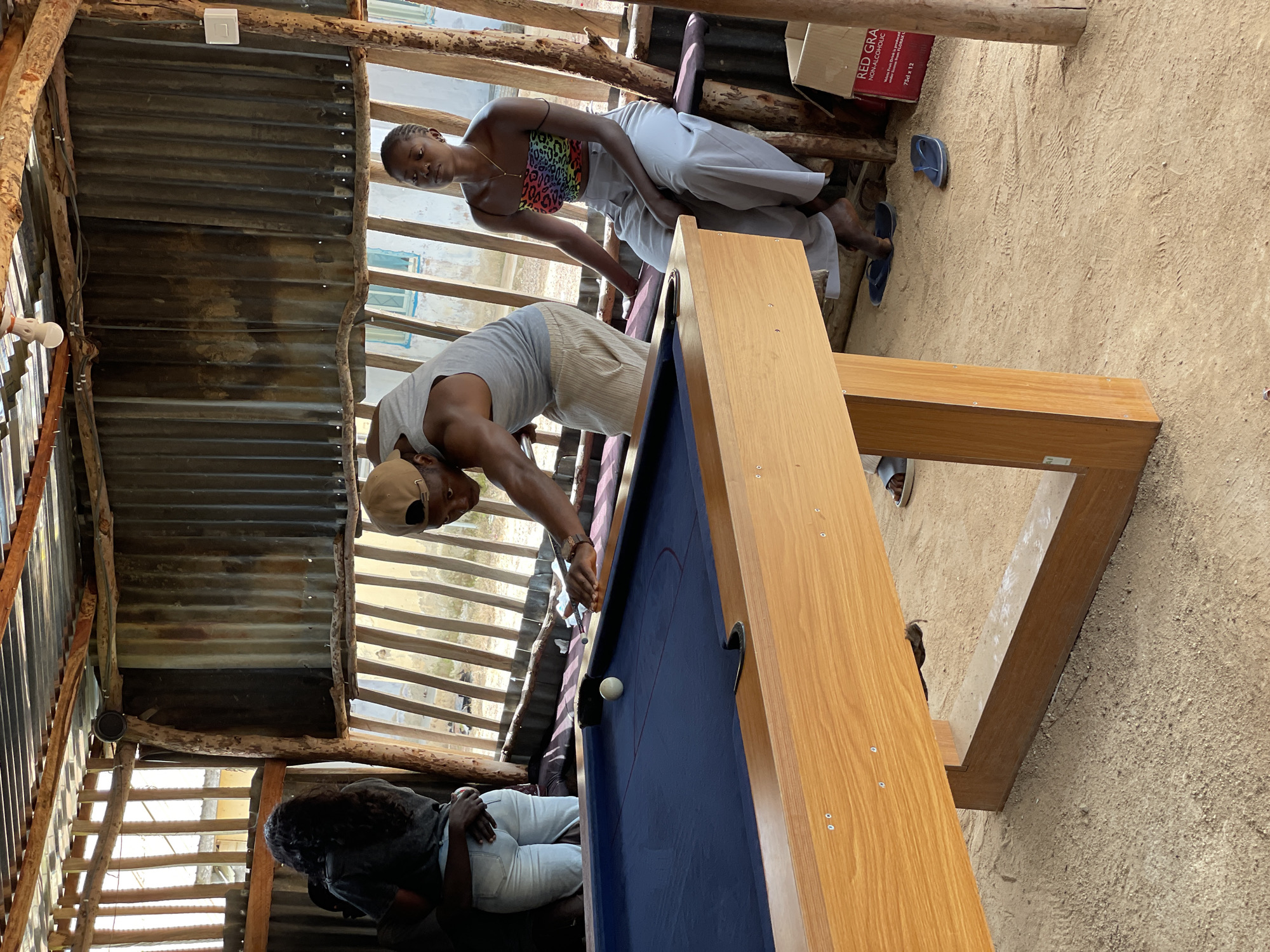 In Durumi, Nigeria, a group of individuals are gathered around a billiards table set up outdoors. The scene is rustic, with a corrugated metal roof supported by wooden beams, and a dirt floor. A man in a gray tank top and a baseball cap is actively engaged in playing billiards, cue in hand, aiming at the white ball. To his right, a woman sits on a wooden bench, wearing a colorful, strapless top and light-colored pants, observing the game. Another woman with long, dark hair is partially visible on the left, her back to the camera, wearing a dark top and light jeans. The billiards table itself is made of wood with a dark blue felt surface. In the background, there are various structures, including what appear to be buildings with windows, and a partially visible cardboard box labeled "RED GRA". The overall atmosphere suggests a casual, community gathering centered around the game.