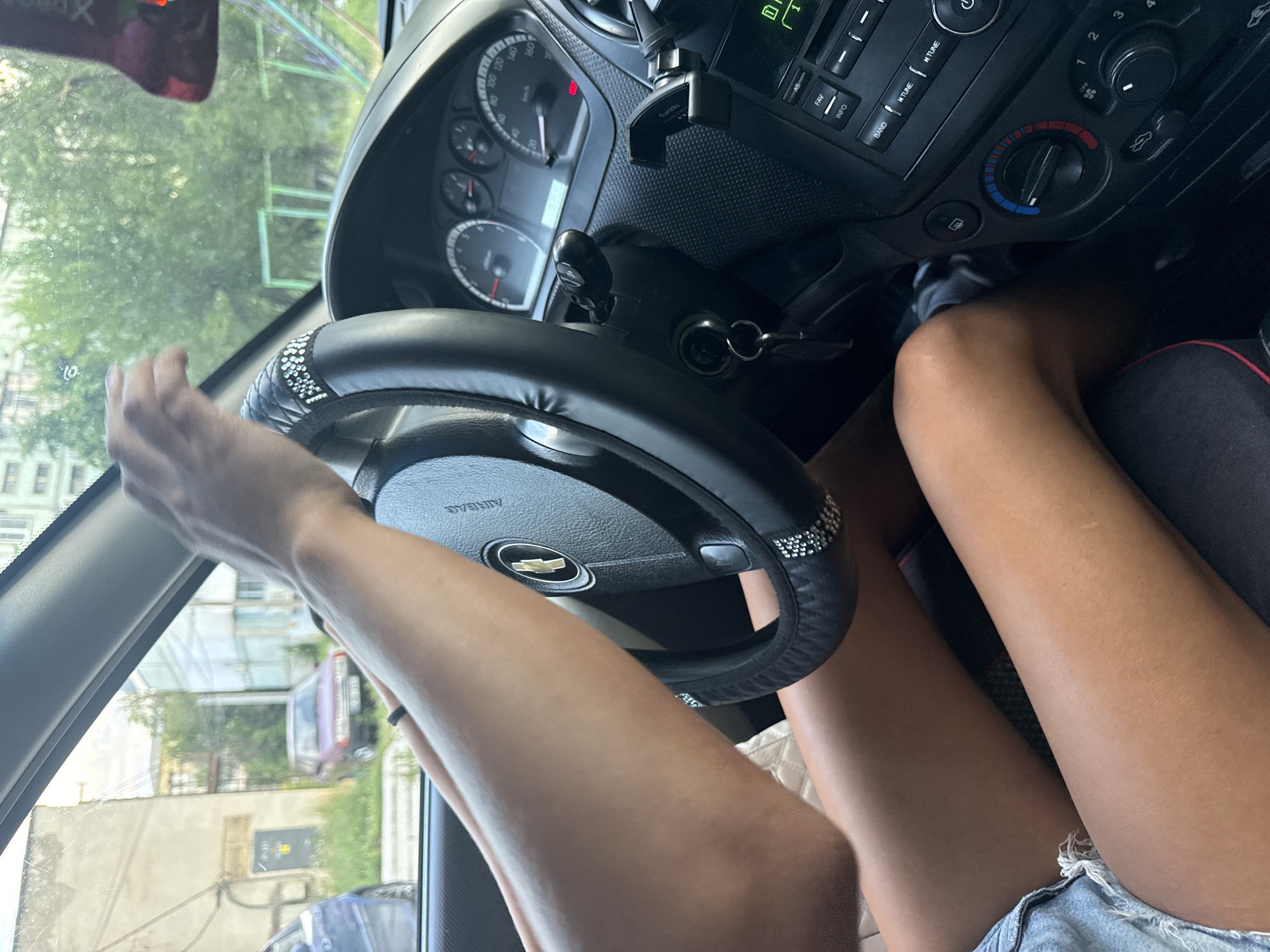The image shows the interior of a car, specifically the dashboard, steering wheel, and a person's legs. The steering wheel is covered in black material with decorative rhinestones around its edge. A car key is visible in the ignition. To the right of the steering wheel are the car's gauges and radio controls. The person's legs are bare and tanned, and they are wearing denim shorts. The exterior of the car is visible through the windows, showing buildings and trees, suggesting an urban or suburban environment. Based on the provided metadata, this scene is located in Basarabeasca, Moldova.