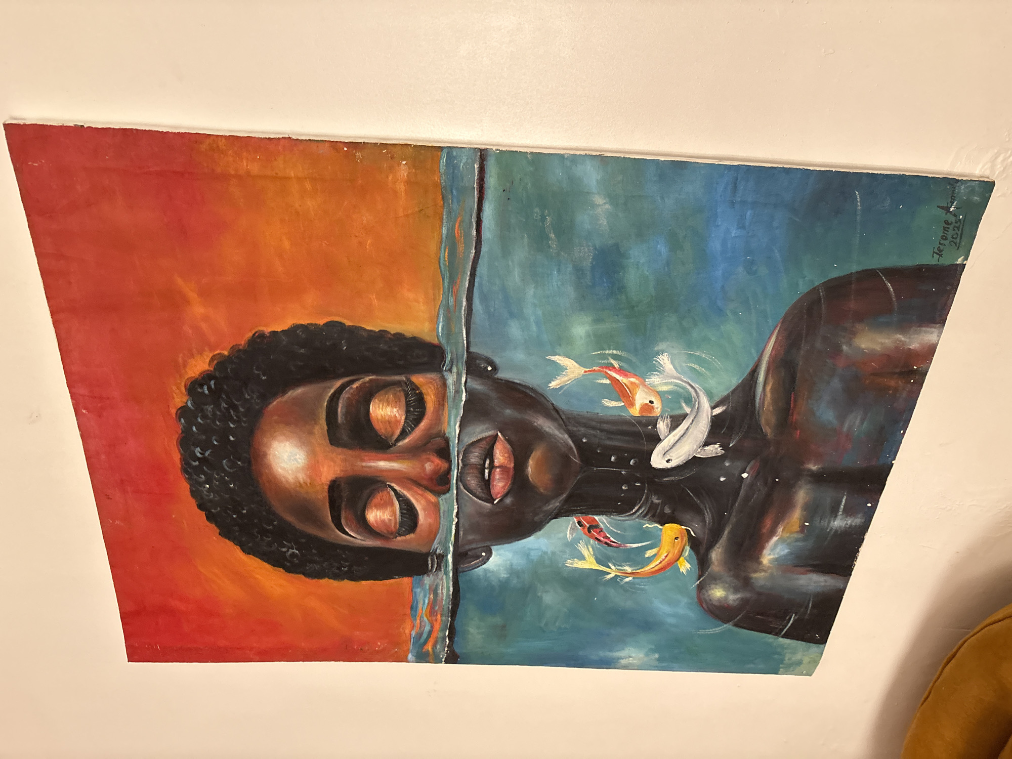 The image shows a painting hanging on a light beige wall. The painting depicts a Black person from the shoulders up, with a red and orange background on the left side of the painting and a blue background on the right side. The face of the person is split in half between the two backgrounds, with closed eyes. The right side of the person's body is painted with darker skin tones, with three koi fish swimming around their head and shoulders. The signature "Jerome April 2023" is visible on the top right corner of the painting. The location, Abuja, Nigeria, is not discernible from the image.