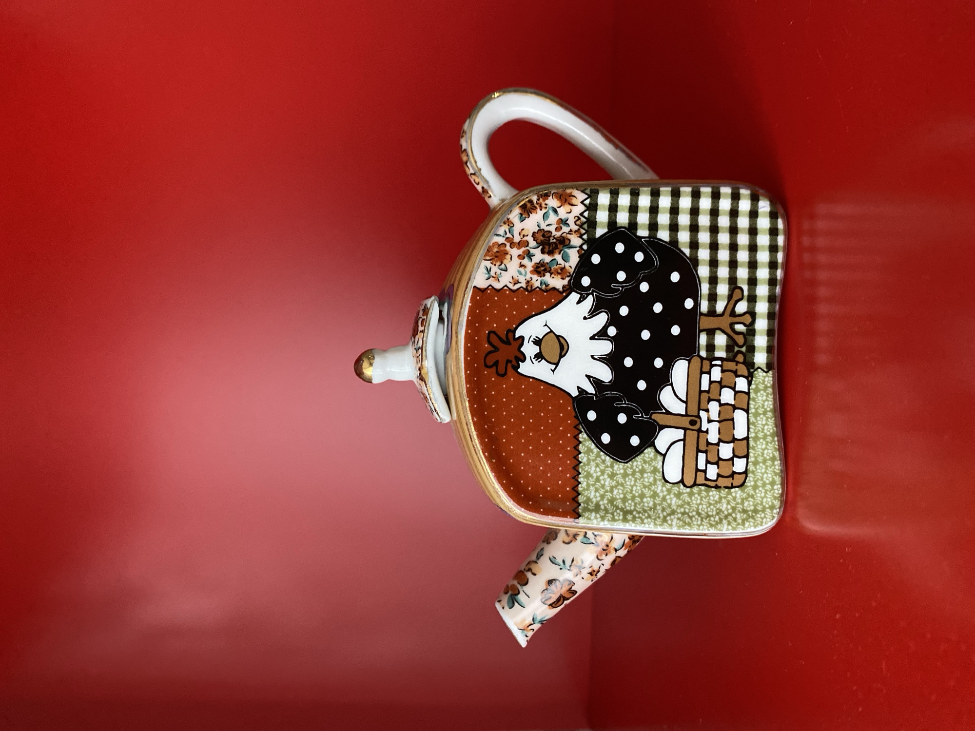 The image shows a ceramic teapot with a decorative design featuring a chicken motif. The teapot has a square body, a handle, and a spout. The design on the teapot incorporates various patterns, including floral, polka dots, gingham, and a chicken with a basket. The teapot is placed on a red surface. The location is not discernible from the image.