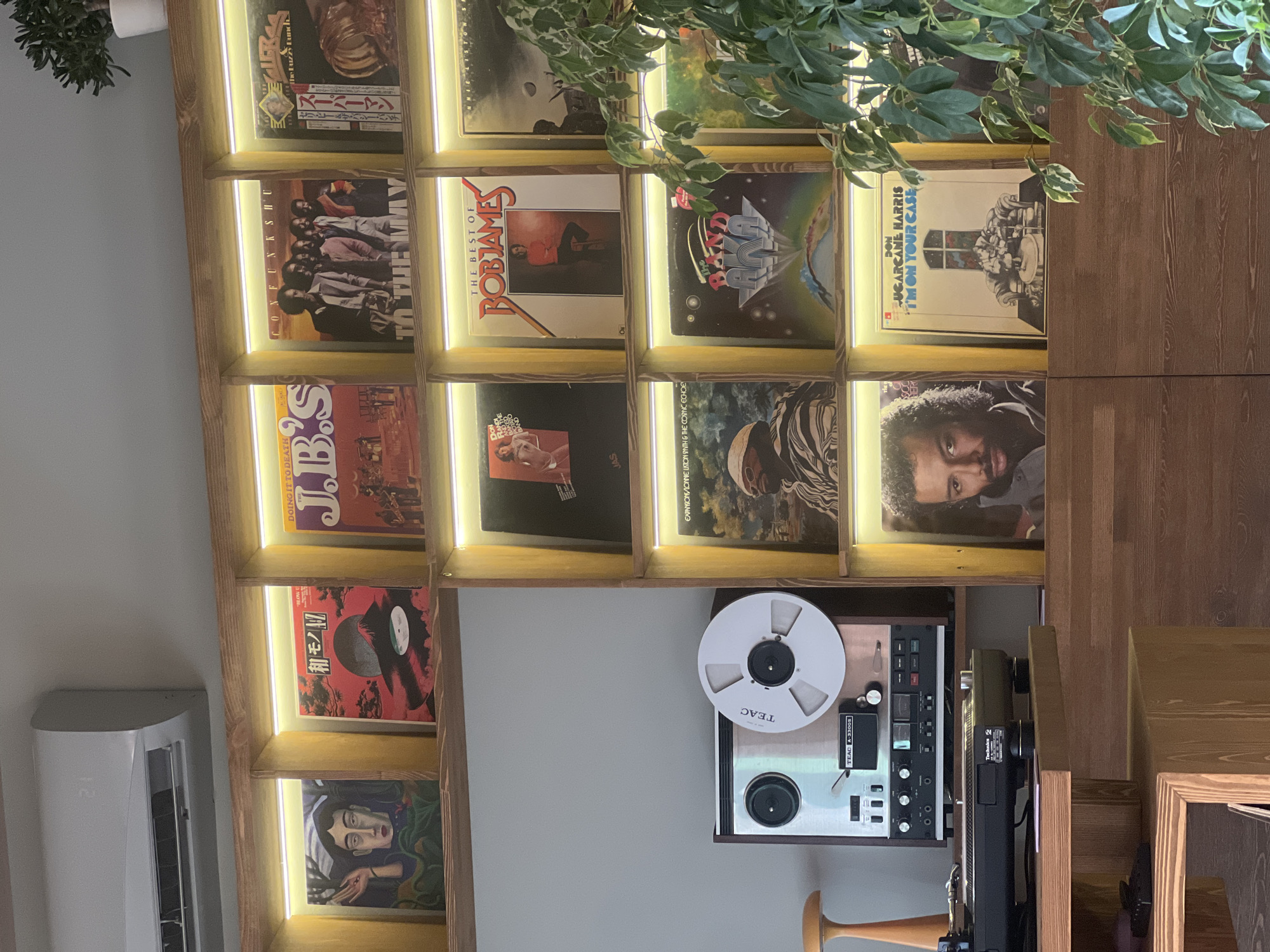 The image shows a shelf filled with vinyl records and a TEAC A-3300S reel-to-reel tape recorder. The records include titles such as "The Best of Bob James," "Con Funk Shun to the May," "J.B.'s Doing It to Death," and "Don 'Sugar Cane' Harris I'm on Your Case." The records and the tape recorder are placed inside a wooden shelving unit that is illuminated by LED lights. There is a plant partially visible at the top, and an air conditioner on the wall to the left. Based on the user prompt history, this scene is likely located in Krasnoyarsk, Russia.