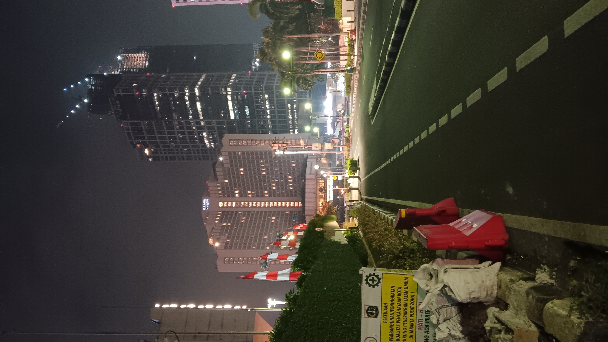 The image depicts a nighttime scene on a street in Central Jakarta, Indonesia, likely in the vicinity of the Grand Hyatt hotel. In the foreground, a dark asphalt road with white dashed lane markings curves to the right. The left side of the road is bordered by a sidewalk with a low concrete barrier. A green hedge runs along the barrier, behind which are several red traffic barriers and piles of white sacks, possibly containing construction debris. A yellow sign with Indonesian text and a cautionary symbol is visible on the left.
Further back, a large, illuminated hotel building with a sign reading "GRAND HYATT" is prominent. To the right of the hotel, a skyscraper under construction is visible, with cranes and scaffolding against the night sky. Other buildings with illuminated windows are also present in the background. Red and white striped flags are mounted on poles along the hedge. Palm trees are scattered throughout the scene, and streetlights illuminate the road. The overall atmosphere is urban and hints at ongoing development. No people are clearly visible in the image.