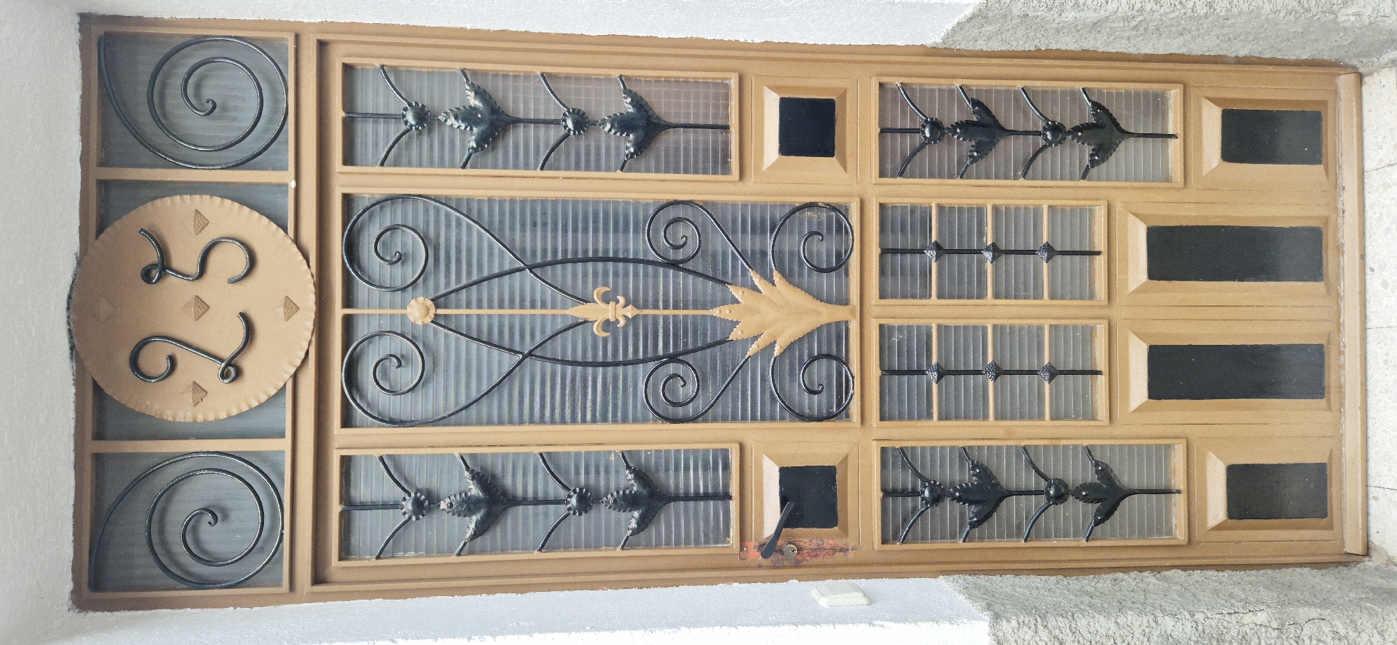 An ornate, weathered wooden door stands out against a textured, light-colored wall. The door features multiple glass panes with decorative wrought-iron elements, including swirling patterns and leaf-like motifs. A circular plaque with the number "25" is prominently displayed at the top of the door. The door has a black handle and a small black mail slot. The lower portion of the door has solid panels with recessed black rectangles. The overall scene suggests an entrance to a residential or commercial building in an urban setting.