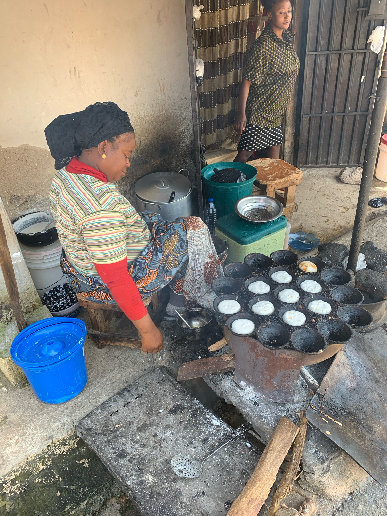 An outdoor scene in Jos, Nigeria, depicts two women engaged in food preparation. A woman in the foreground, seated on a wooden stool, wears a black headscarf, a striped shirt with a red collar, and a patterned skirt. She is actively cooking, using a traditional charcoal stove with a round, sectional pan to fry small, white pastries. Beside her, to the left, is a large blue plastic bucket. In the background, another woman stands near a doorway, looking towards the viewer. She is dressed in a patterned shirt and a polka-dot skirt. The environment suggests a modest, functional outdoor cooking area with concrete ground, cooking implements, and various containers.