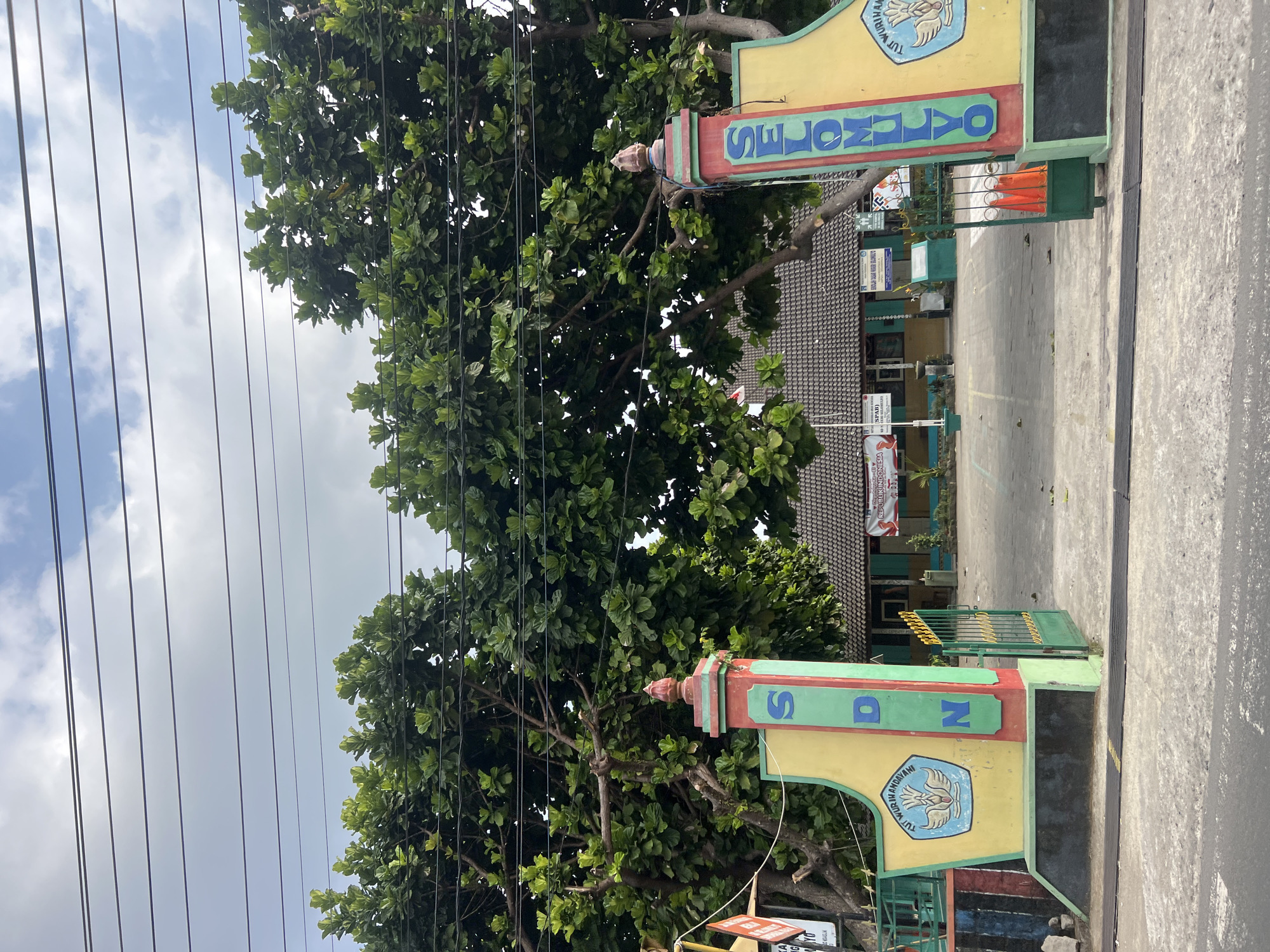 The image shows the entrance to SDN Selomulyo, a primary school located in Ngaglik, Indonesia. The entrance features two large pillars, one with the text "SDN" and the other with "SELOMULYO" in vertical lettering. Both pillars are decorated with colorful designs and feature the Indonesian national emblem on the left pillar. A green metal gate is open, leading into the school grounds. Behind the entrance, a school building with a tiled roof is visible, along with trees and some banners. The sky is partly cloudy, and power lines are visible across the top of the image. The scene appears to be a typical school entrance on a bright day.