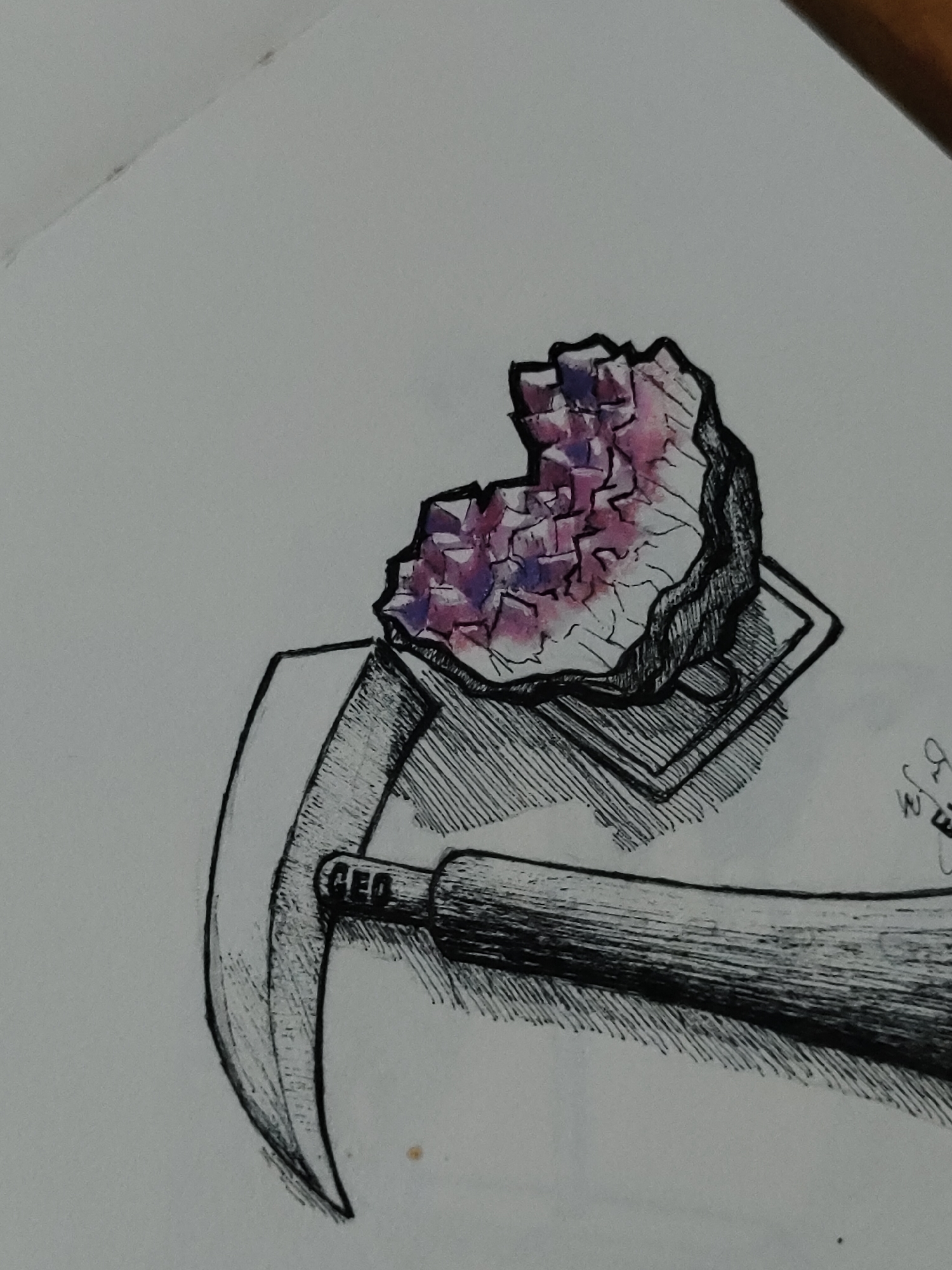 An overhead photograph displays a hand-drawn illustration rendered primarily in black ink with partial color accents, set against a white paper surface in an unspecified indoor environment. The drawing depicts a geode or crystal cluster positioned above a geological pickaxe.
The geode, located in the upper-middle section of the frame, is outlined in thick black ink and features a rough, irregular exterior. Its interior reveals numerous faceted crystal formations, colored in varying shades of pink and purple, with some areas appearing more blue-violet. Fine black lines are used for texture and shading on the geode's outer rock casing. This geode rests on a multi-layered, rectangular base, rendered with dense cross-hatching for shading.
Beneath and extending from the bottom-left to the middle-right, a geological pickaxe is illustrated. The handle is thick and cylindrical, detailed with extensive horizontal cross-hatching. The letters "GEO" are discernible in black ink on the handle near its junction with the pickaxe head. The head of the tool is curved and tapers to a point on the left, also shaded with cross-hatching to indicate its metallic texture and form.
The white paper exhibits a faint diagonal crease or fold line in the upper-left corner. Small, indistinct specks or marks are present on the paper surface. A partial signature or initial, appearing to be "BJM," is visible in the lower-right quadrant of the drawing.