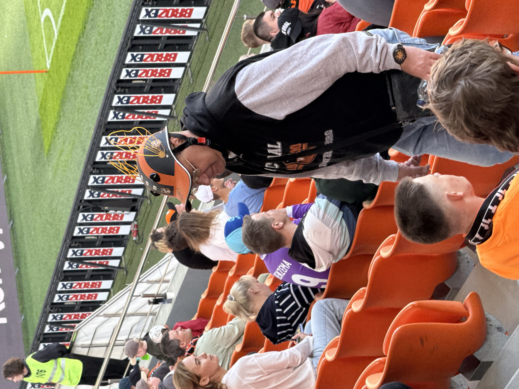 The image shows a crowd of people in orange stadium seating, likely at a sporting event. A soccer field with white boundary lines is visible to the side of the image. Some of the crowd are watching the game. One man is wearing an orange helmet. There is a person wearing a yellow and black vest. There is signage with "BROZEX" printed on it. Some printed text with a logo on a man's clothing contains "IC VAL BITEL ARG" near a logo. It is likely the Yekaterinburg Arena, in Yekaterinburg, Russia.