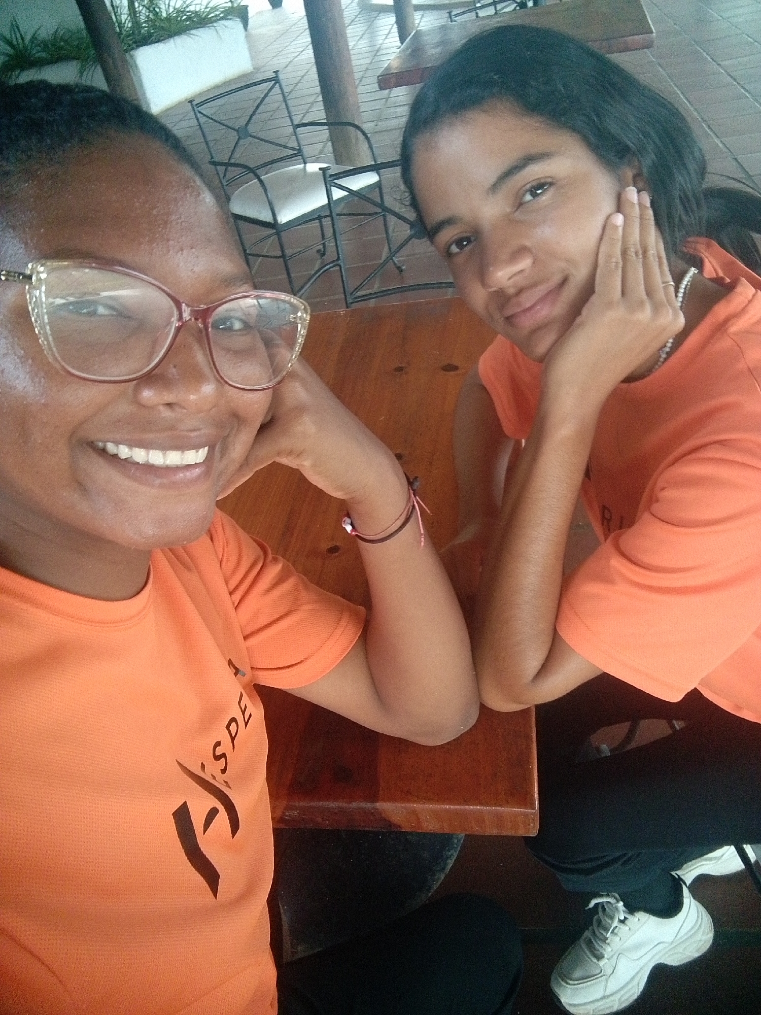 Two young women pose for a selfie at a wooden table. The woman on the left wears glasses and smiles broadly. Both women are wearing orange t-shirts with black writing. They both rest their heads on their hands, leaning on the table. One wears white sneakers. The setting appears to be an outdoor seating area of a restaurant or cafe, with wrought iron chairs and potted plants visible in the background. This photo was taken in Pedro González, Venezuela.