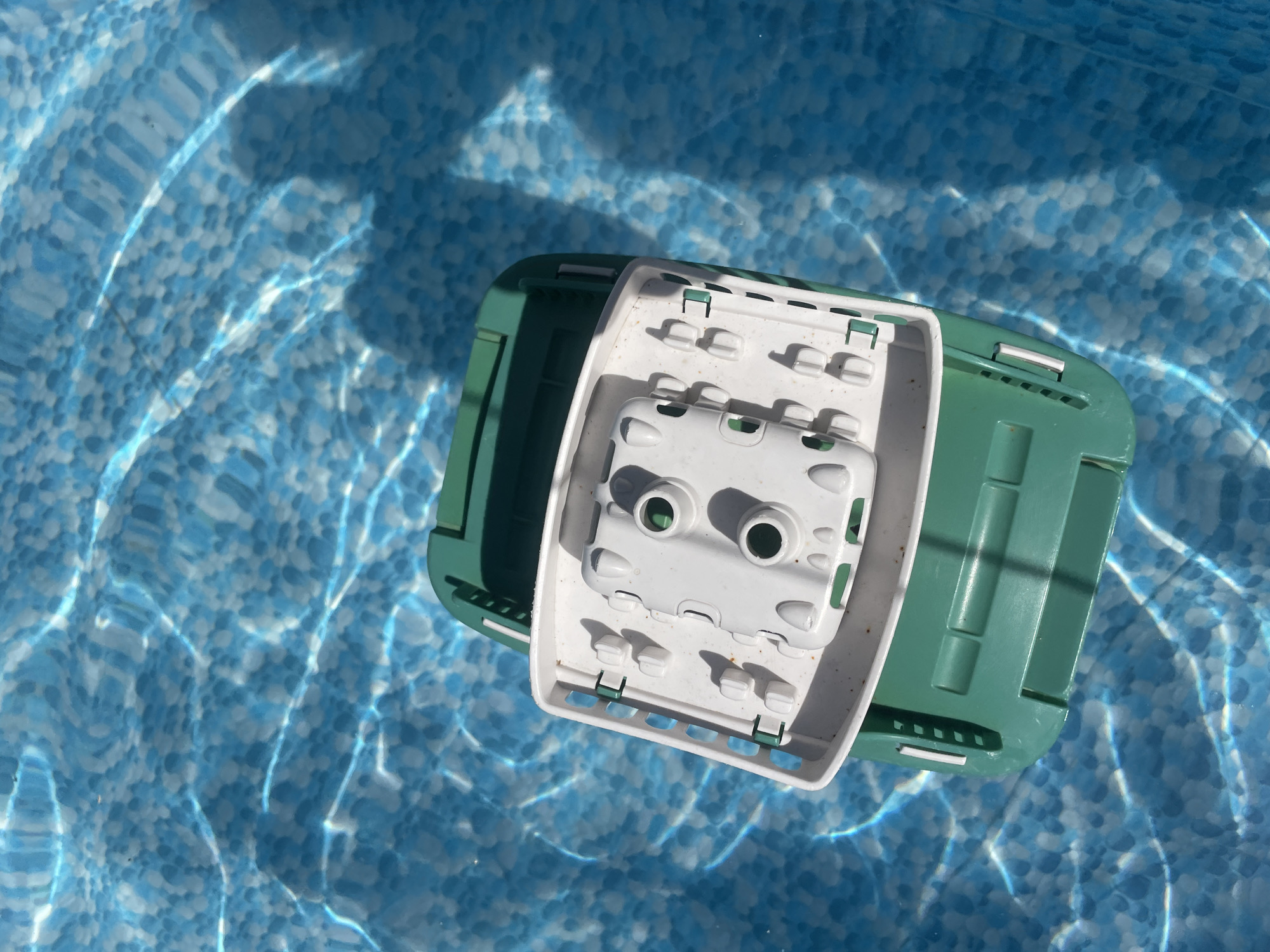 An overhead shot captures a green and white plastic toy boat floating in clear blue water, likely a swimming pool, due to the patterned bottom and refracted light. The toy has a green hull and a white upper deck with molded seating and two circular openings. Sunlight casts wavy patterns of light and shadow on the water's surface and the pool floor. The scene is static, with no people or discernible action. The toy boat is the sole subject. The location is identified as Baranovka, Russia.