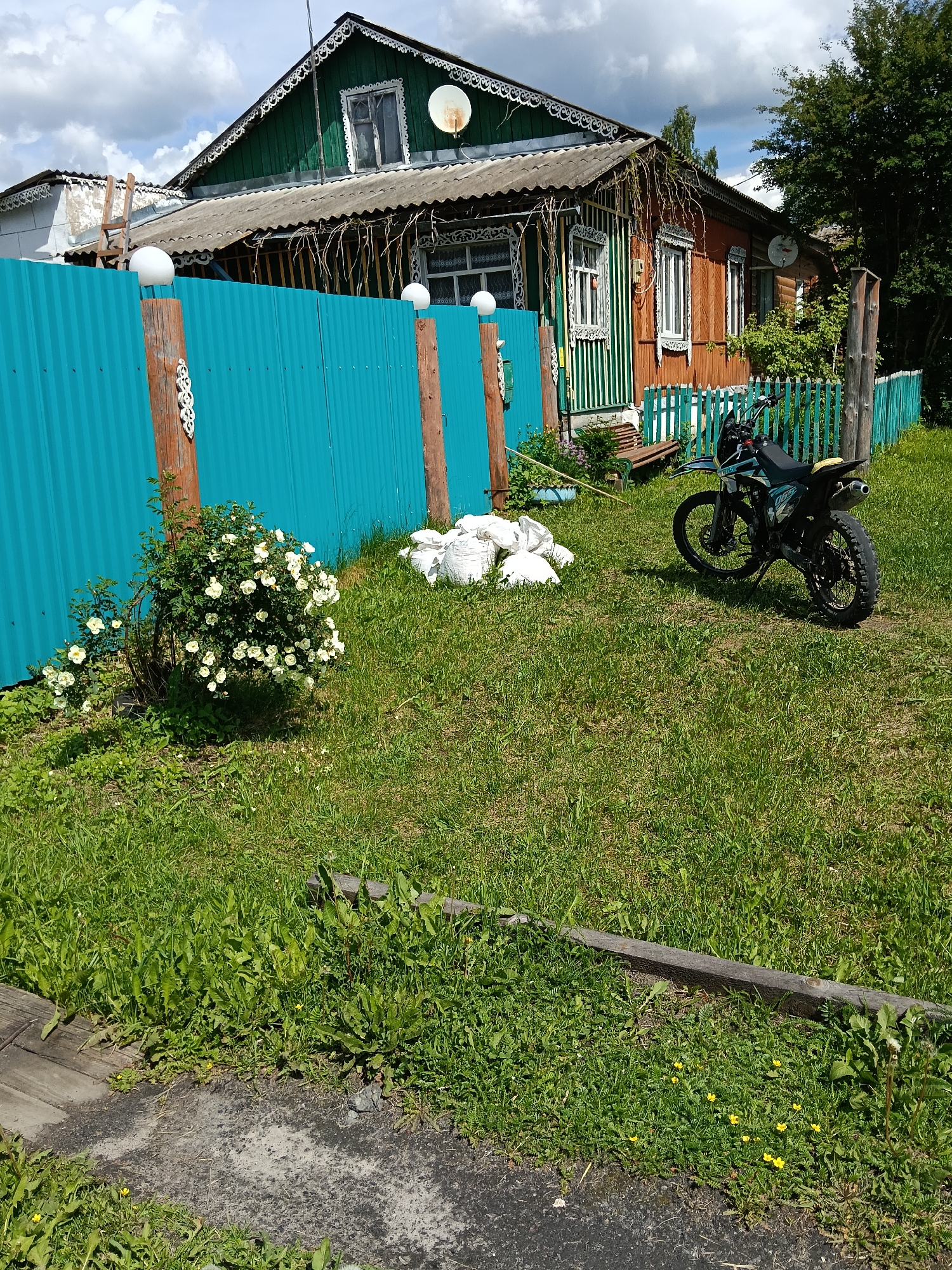 This image depicts a rural or semi-rural residential scene in Красный Маяк, Russia, featuring a traditional wooden house and its yard under a bright, partly cloudy sky.
The prominent two-story house, built from wood, exhibits intricate decorative white trim, often referred to as "gingerbread" style, around its windows and roofline. The upper section of the house is painted dark green, while the lower part is a warm reddish-brown. It has a corrugated gray roof and two white satellite dishes attached to its exterior walls. A wooden ladder leans against a side section of the house.
In front of the house, a bright turquoise corrugated metal fence with wooden posts defines the property boundary. Some posts are adorned with white spherical light fixtures on top, and one post features a white, decorative carved element. Inside the yard, to the left of the fence, a lush rose bush with numerous white or very light yellow flowers is blooming.
Further into the yard, several white sacks are piled on the grass, possibly containing gardening materials or waste. A black dirt bike or motorcycle with yellow accents is parked on the grass to the right. A smaller, pale green wooden fence is visible behind the motorcycle, bordering another section of the property. The ground is covered with vibrant green grass, interspersed with weeds and some small yellow wildflowers. Two wooden planks lie on the ground in the foreground, near a patch of bare soil.
The time of day is daytime, evidenced by the clear shadows cast by the objects and the bright illumination from the sky, which shows some visible clouds. No people are visible, and there is no legible text on any of the objects, though the motorcycle has some unreadable branding.