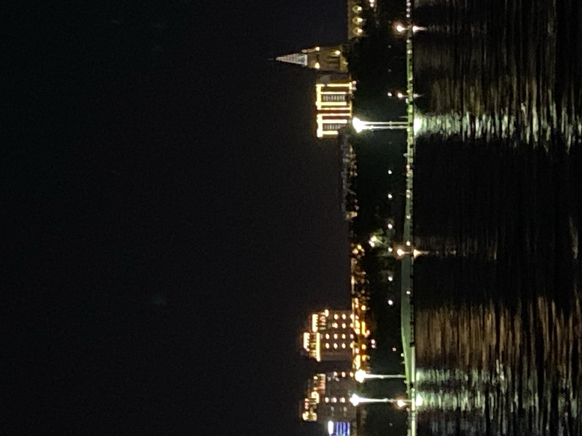 This is a nighttime photograph of a cityscape with a body of water in the foreground. The water reflects the lights from the buildings. In the background, several buildings are visible. To the left, there are two illuminated multi-story buildings. In the center, there is a structure with bright yellow lights that form a rectangular pattern, and to its right, a tall, steeple-like building with lights. The sky is dark, indicating nighttime. The image appears to be taken from across the water, possibly from a boat or the opposite shore. The lights and reflections suggest an urban environment. Given the context provided that the image is of Blagoveshchensk, Russia, it is likely a view of the Amur River with buildings on the embankment. There are no people visible in the image.