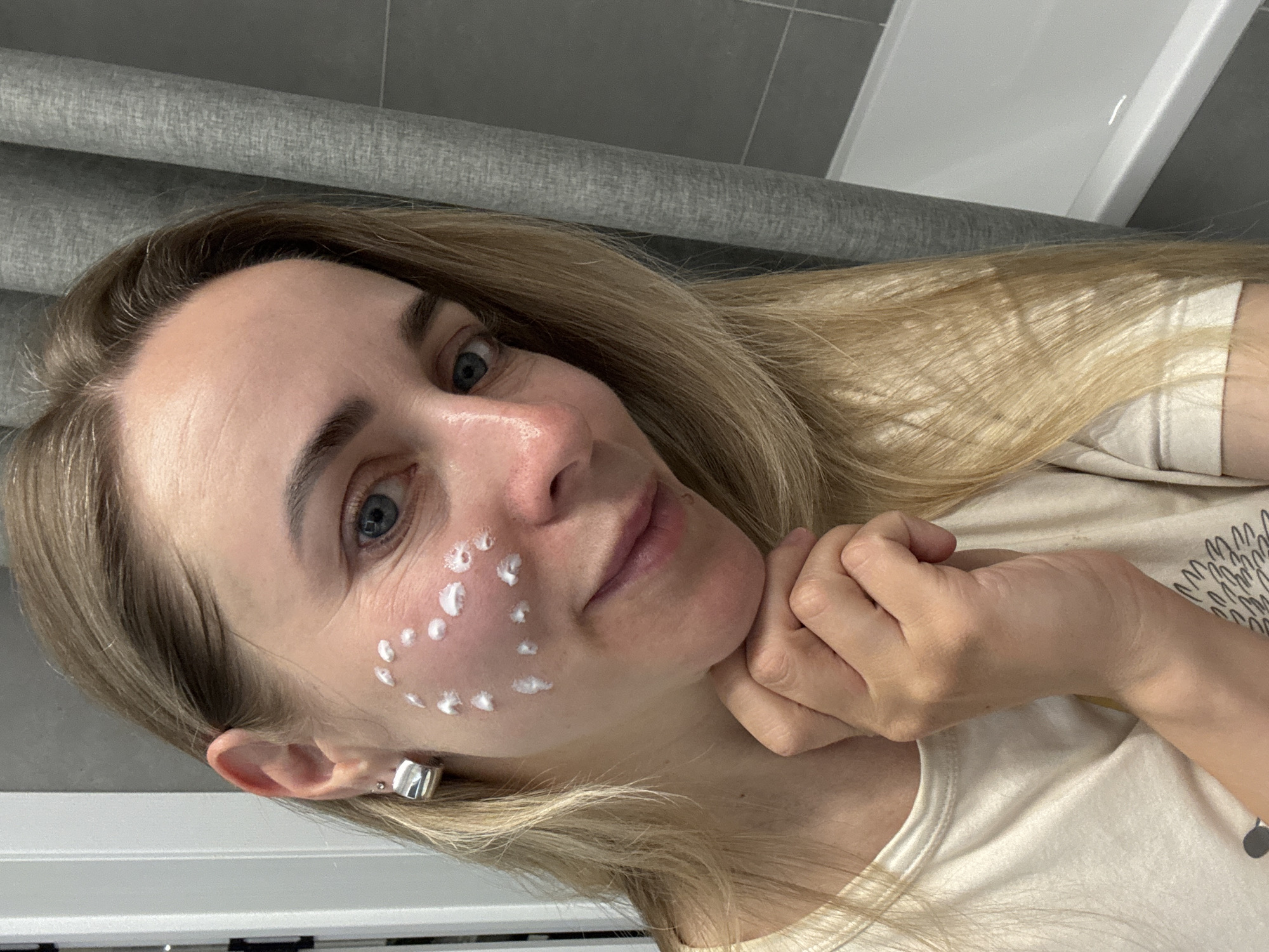 A blonde woman is positioned at an oblique angle to the camera, with her face turned towards the lens. She has fair skin, blue eyes, and wrinkles are visible on her forehead. Multiple small dollops of a white substance are visible on her left cheek. Her right hand is positioned beneath her chin. She wears a white t-shirt, and a silver earring is visible in her left ear. The background consists of a grey tiled wall and a white object, possibly a bathtub or sink. There is no indication that this image was taken in Chaikovsky, Russia.