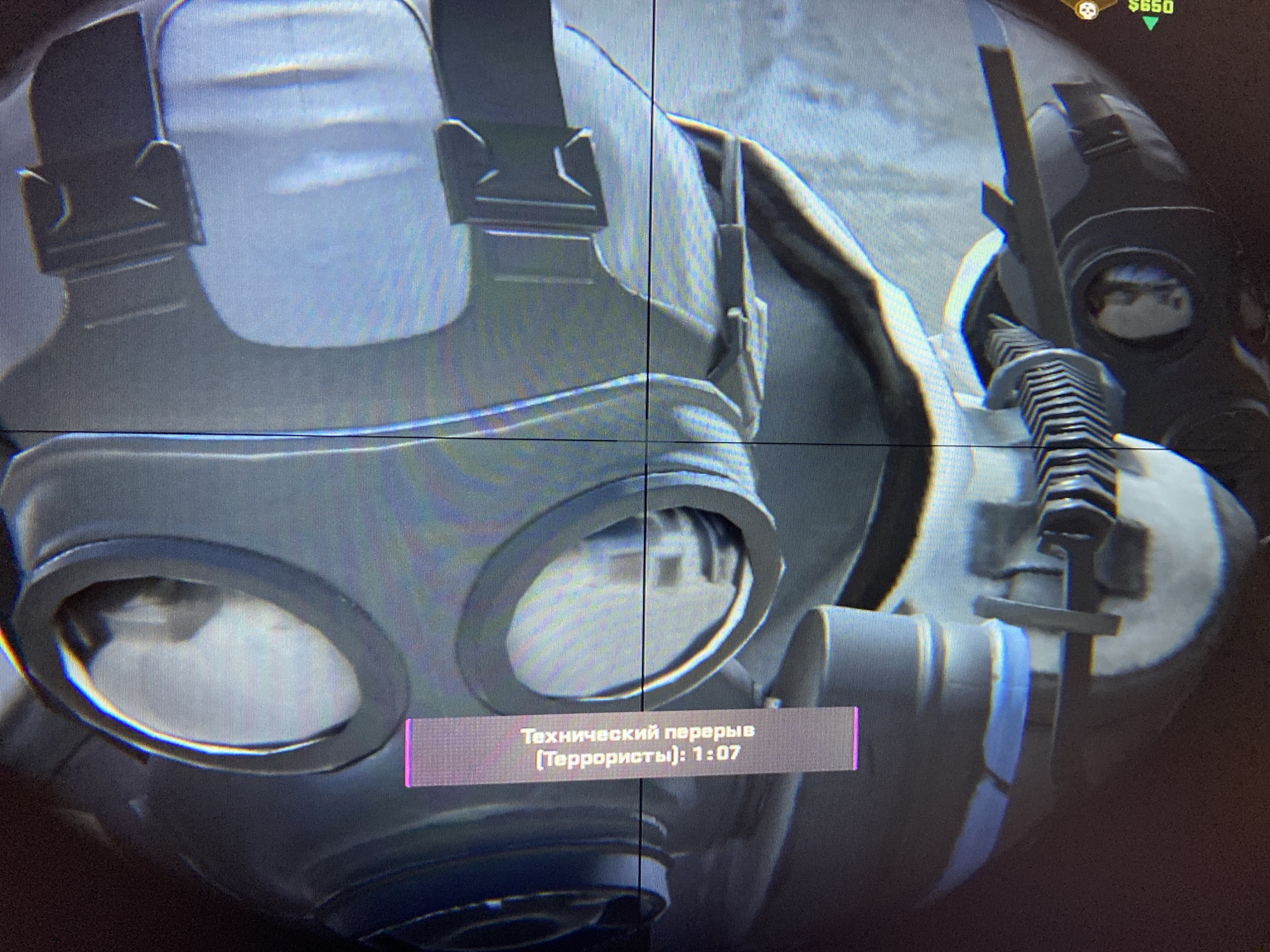 The image displays a close-up, screen-captured view of a video game, observed through a prominent crosshair reticle. The central subject is a character wearing a grey gas mask with two large, reflective eye-pieces, and part of a tactical vest or harness with visible buckles. To the right, a section of a weapon, possibly with a textured grip and a blade-like attachment, is visible. The background is indistinct and blurred, typical of a digital game environment.
In the top right corner of the screen, a small icon of a skull is displayed alongside "$650," likely indicating in-game currency or score. A translucent, purple-pink rectangular overlay appears in the lower center of the screen, containing white Russian text. This text reads "Технический перерыв (Террористы): 1:07," which translates to "Technical pause (Terrorists): 1:07." This suggests a temporary halt in gameplay, possibly in a competitive match.
The image itself is a photograph of a digital screen, evident from the visible pixel grid. The lighting is bright, reflecting off the gas mask lenses. No specific time of day, weather, or discernible location cues within the game are visible, other than the general context of a tactical shooter. There are no visual elements within the image to indicate the provided location context of Yerevan, Armenia.