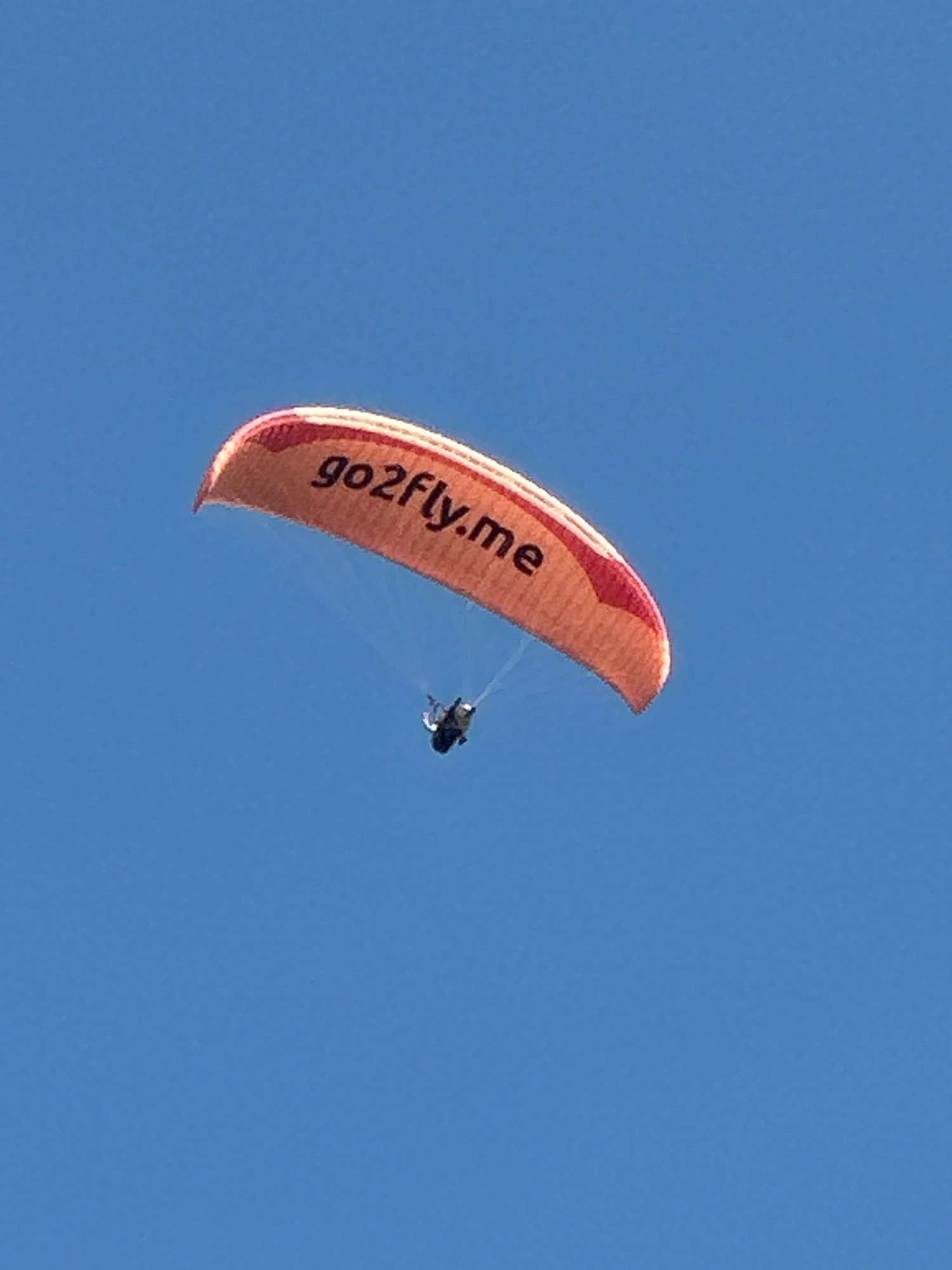 A paraglider with an orange canopy emblazoned with the text "go2fly.me" is captured in mid-flight against a clear blue sky. The paraglider's harness is visible, carrying a person dressed in dark clothing. The scene appears to be set in Boreti, Montenegro, a coastal town known for its scenic beauty and potential for recreational activities. The image focuses on the paraglider, with the vast expanse of the sky dominating the background. There are no other objects or people prominently featured in the frame, emphasizing the solitary nature of the paraglider's flight. The lighting suggests it is daytime, with sunlight illuminating the paraglider and canopy.