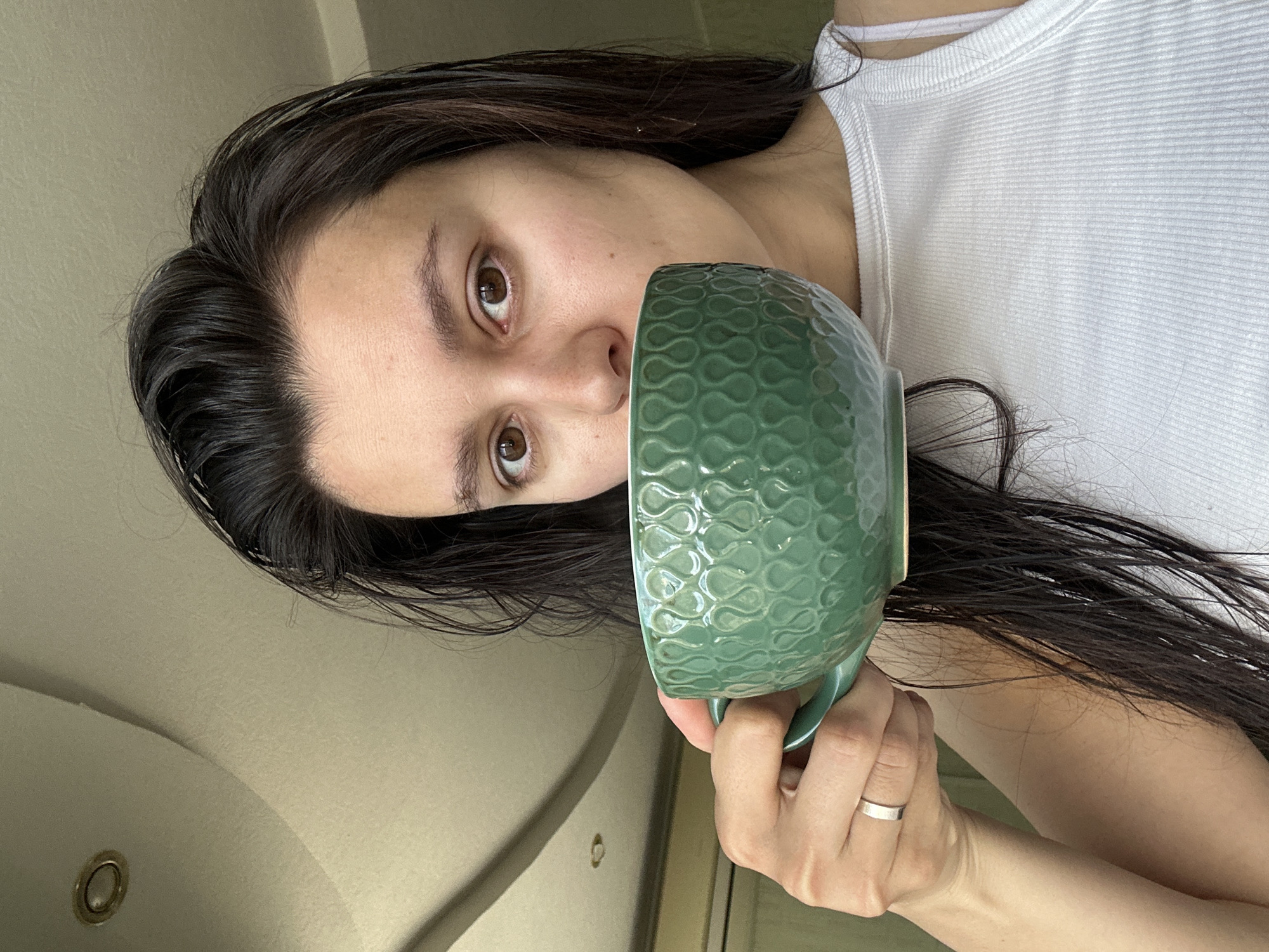 A young woman with dark, wet hair and brown eyes is holding a green mug with a textured, wave-like pattern to her mouth. She is wearing a white ribbed tank top. A silver band is visible on her left ring finger. The background includes a beige wall and a curved ceiling with a recessed light. The image appears to be taken indoors, possibly in a bathroom. There is no information available to determine if the image was taken in Chaikovsky, Russia.