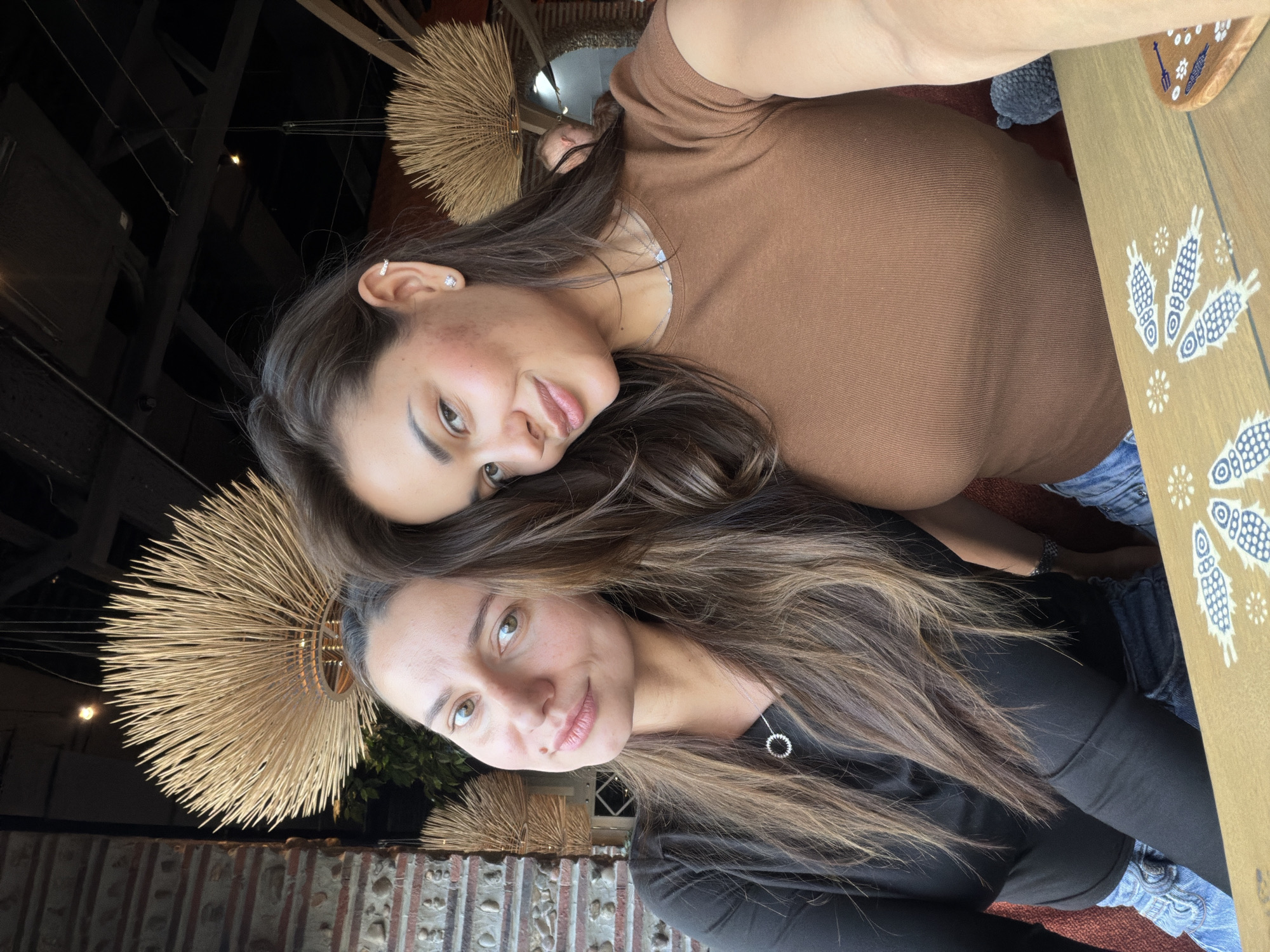 Two women are posing for a selfie. The woman on top is wearing a brown long-sleeved shirt and jeans. The woman on the bottom is wearing a black long-sleeved shirt and jeans. Both women have long, brown hair. They are sitting at a table with a patterned cloth on it. The background includes a textured wall, light fixtures resembling sunbursts, and glimpses of the outdoor environment. The location is inferred to be in Krasnoyarsk, Russia based on the presence of people and general environment, but I cannot verify this for certain.