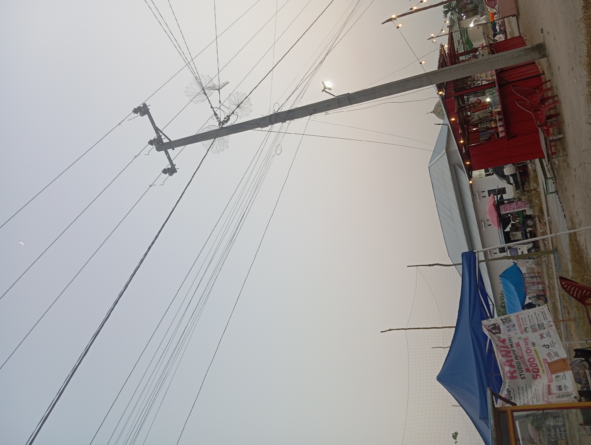 The image depicts an outdoor scene with a prominent utility pole supporting numerous power lines. The sky is overcast and hazy, suggesting either dawn, dusk, or a dusty atmosphere. In the foreground, there are several stalls or small shops. One stall, to the left, is covered by a blue canopy and features a banner advertising a "Rank Studio Photo Mini" with a price of "5000/Orang" (5000 per person), indicating a photography service. Adjacent to this is a red stall, possibly a food or drink vendor, with a counter displaying various items and illuminated by string lights. Red chairs are placed in front of the red stall. In the background, a building with a metal roof is visible, and further in the distance, more structures and greenery can be seen. The ground appears to be dirt or sand. The presence of the stalls and the general ambiance suggest a market or an outdoor event in a semi-rural or suburban area. Based on the location provided, this scene is in the city of Way Dente, Indonesia.