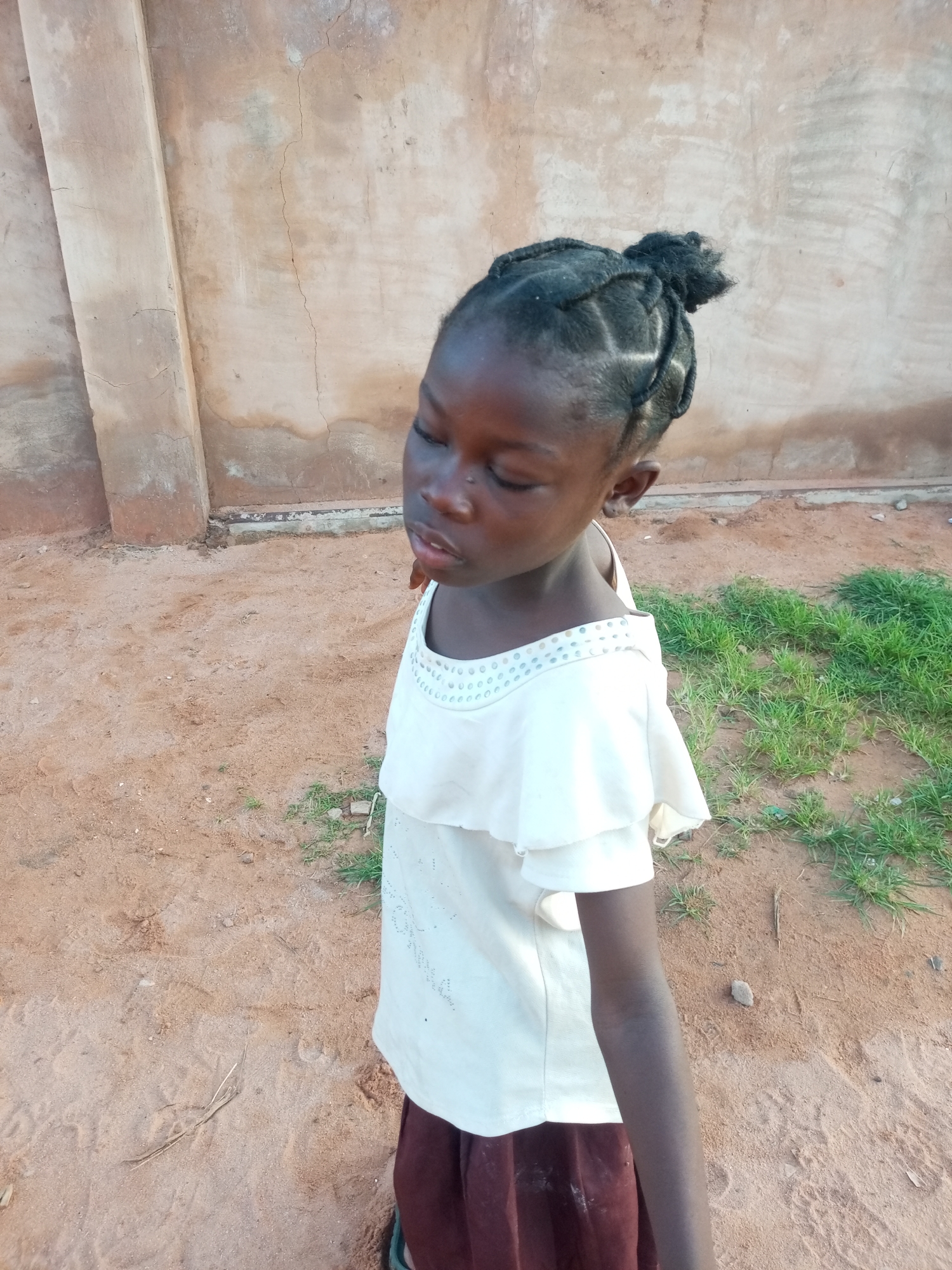 A young Black girl is standing outdoors, looking down and slightly to her left. Her hair is styled in braids and pulled into a bun. She is wearing a white, ruffled shirt with decorative beadwork around the neckline and a dark reddish-brown skirt. The ground beneath her is composed of loose sand with patches of green grass and some scattered small stones. Behind her is a textured, light brown wall, possibly made of cement or stucco, with vertical architectural elements. The overall setting appears to be a simple outdoor environment, potentially in a rural or less developed urban area, consistent with locations in Tempure, Nigeria.
