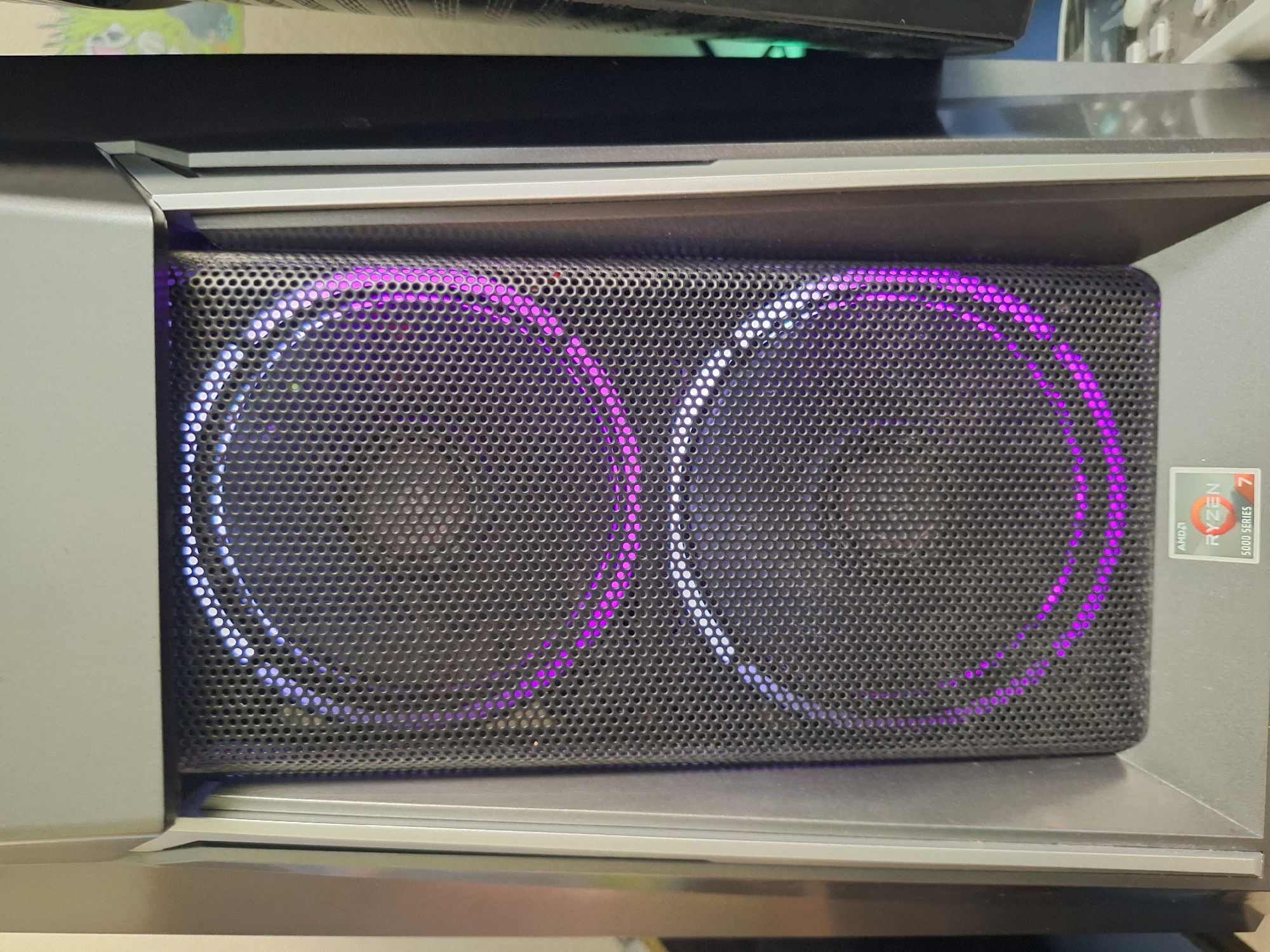 The image shows the front of a computer tower case with two large, circular fans visible behind a perforated metal grille. The fans are illuminated with purple and white LED lights, creating a glowing effect. In the lower right corner of the computer case, there is a sticker indicating "AMD RYZEN 5000 SERIES 7". The computer case is dark gray and appears to be in a home or office environment. There is no indication of the location being Varna, Bulgaria, or any specific city.