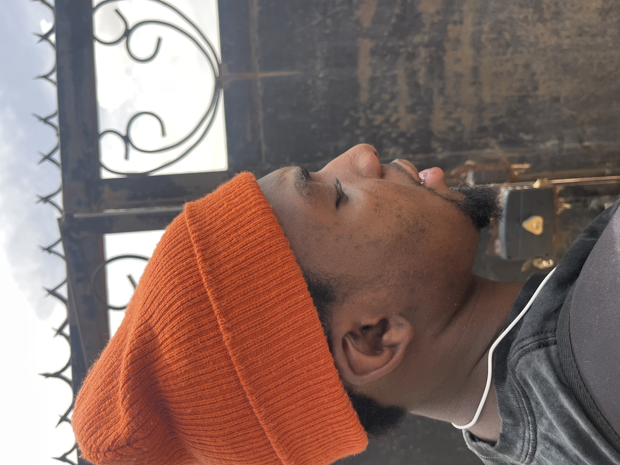 A man wearing an orange beanie and a black shirt is standing in front of a weathered gate with ornate metalwork in Jos, Nigeria. The gate, constructed from dark metal, features decorative swirls and sharp points along the top edge. The man's head is tilted upwards, his eyes are closed. A white cord, possibly headphones, is visible around his neck. The background is blurred.