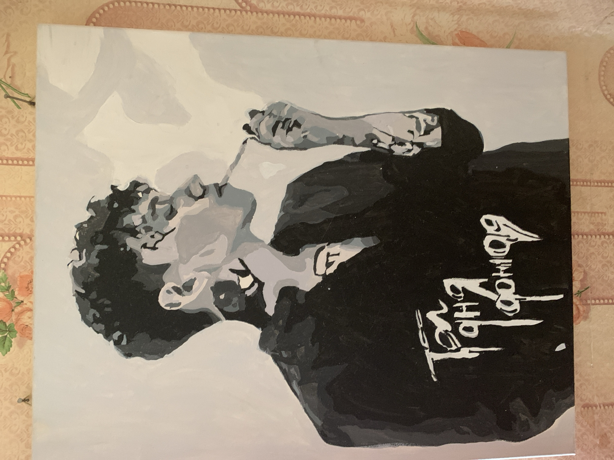 This is a painting of a person in a grayscale style. The subject is shown from the chest up, with their head tilted back and eyes closed. They are holding a cigarette or similar object to their mouth. The person is wearing a black shirt with white text on it. The background is a light gray color, with some abstract shapes in a lighter shade of gray. The overall mood of the painting is somber or introspective.
The visible text on the shirt appears to be in a stylized script. While the exact words are difficult to decipher due to the artistic rendering, it has a calligraphic quality.
The painting is placed on a surface that appears to be a table with a textured, patterned tablecloth featuring floral motifs. The tablecloth has a warm, peachy-pink tone with hints of green and brown. The setting does not provide any specific location cues beyond it being indoors. There are no indications of the time of day or weather conditions.