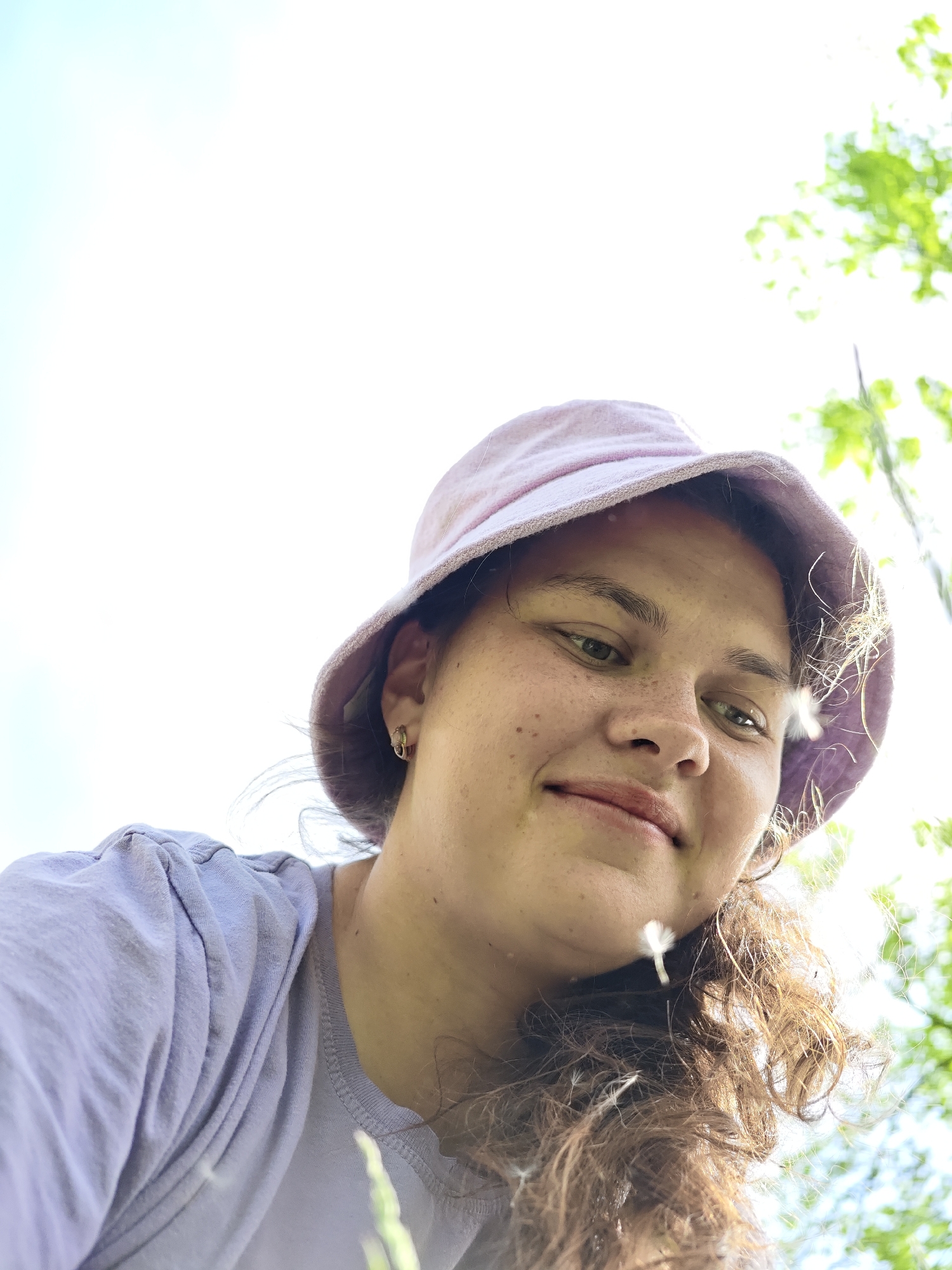 A low-angle, medium shot depicts a young woman with freckles and curly brown hair, partially obscured by the camera angle. She wears a light purple bucket hat and a matching short-sleeved shirt. A small gold earring is visible in her left ear. Several dandelion seeds are floating around her face and hair. The background is a blurry mix of bright sky and green foliage. No information is available to confirm that the image was taken in the city of Chaikovsky, Russia.