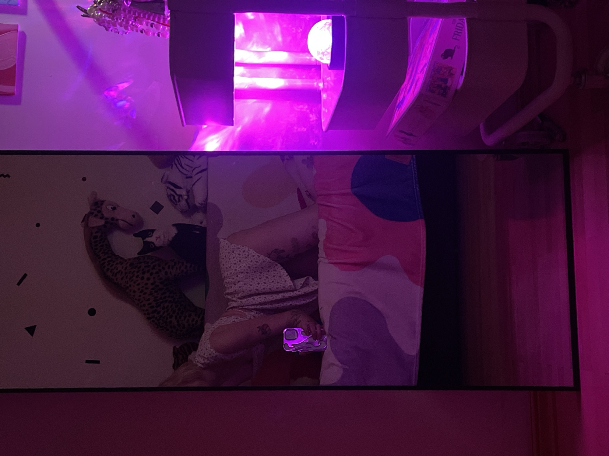 This image captures a mirror selfie taken indoors, likely in a bedroom or living space in Belgrade, Serbia. The scene is bathed in a vibrant purple light, emanating from an artificial light source, possibly LED strips or a lamp, positioned above the mirror.
In the mirror's reflection, a person is seen lying down on what appears to be a bed or couch, wearing light-colored, patterned pajamas. Visible tattoos are on their limbs. They are holding a smartphone encased in a distinctive purple, flame-patterned phone case. Next to the person are a large leopard-print stuffed animal, possibly a giraffe, and a smaller black-and-white cat-like plush toy. The bedding features abstract patterns in shades of pink, purple, and white. The wall in the reflection is adorned with black geometric shapes, including dashes, triangles, and circles.
Above the mirror, in the actual room, the intense purple light illuminates a white shelf or structure. On this shelf, a book or box is visible with the text "FRIDA" and the silhouette of an animal, possibly a dinosaur or lizard. Portions of the word "FRIDAY" might also be present. To the left of the light source, a small piece of wall art with pink and white abstract shapes can be seen. A decorative, possibly crystal, light fixture or chandelier hangs on the wall nearby. To the right, a white metal frame, likely part of furniture, is visible. The overall ambiance is relaxed and suggests an evening or nighttime setting due to the prominent artificial lighting.