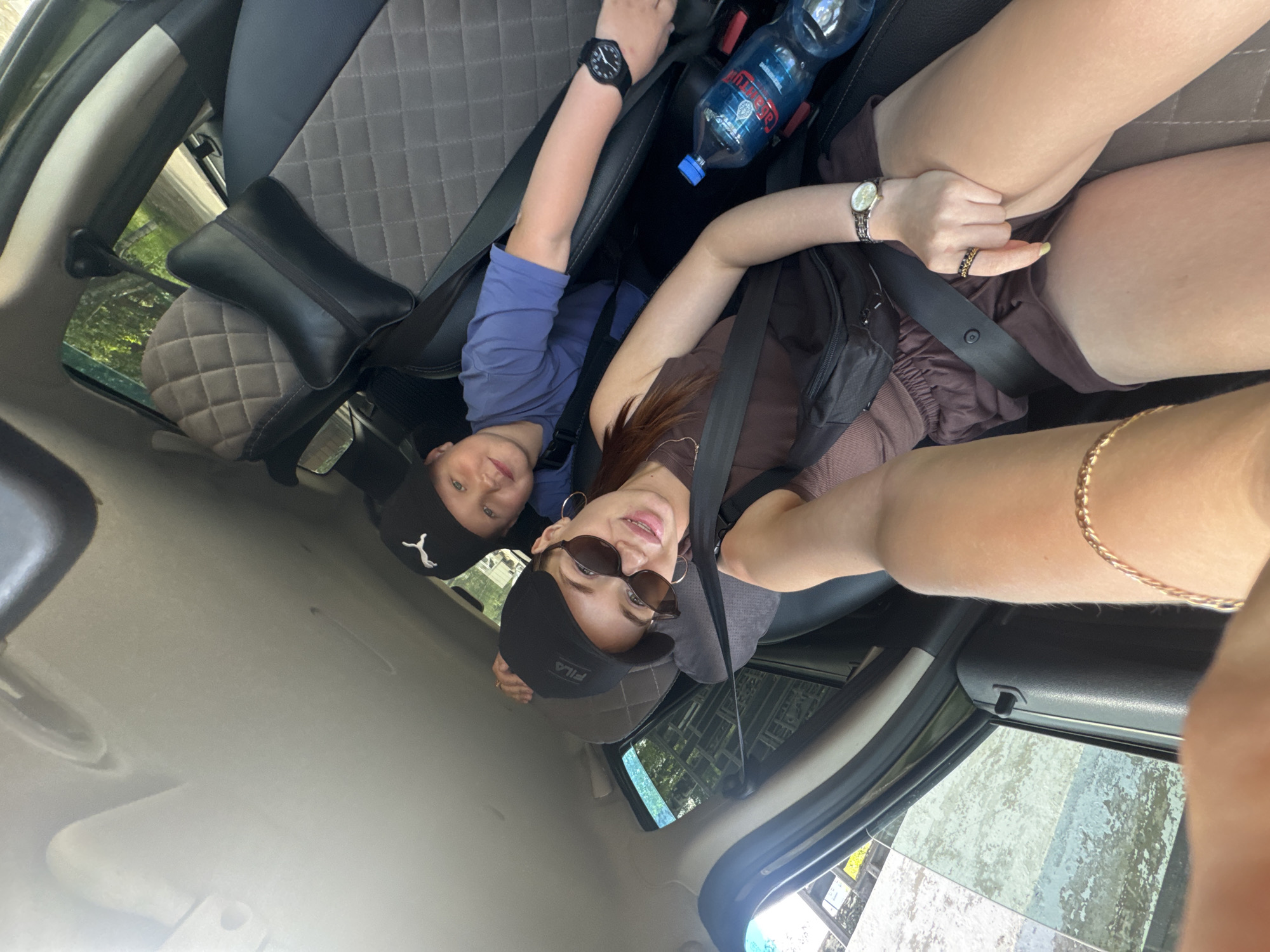 In a vehicle, two individuals, a woman and a child, are seated. The woman, in the foreground, is wearing a brown top, shorts, sunglasses, and a black baseball cap. She has a watch on her wrist and is holding a phone, presumably taking the photo. The child, in the background, is wearing a blue t-shirt and a black baseball cap. A bottle of water is visible on the seat next to the child. The interior of the car appears to be a standard passenger vehicle. The presence of trees and greenery visible through the windows suggests the vehicle is in motion, possibly on a road outside of urban infrastructure, making a specific location within the city of Chaikovsky, Russia, difficult to ascertain.