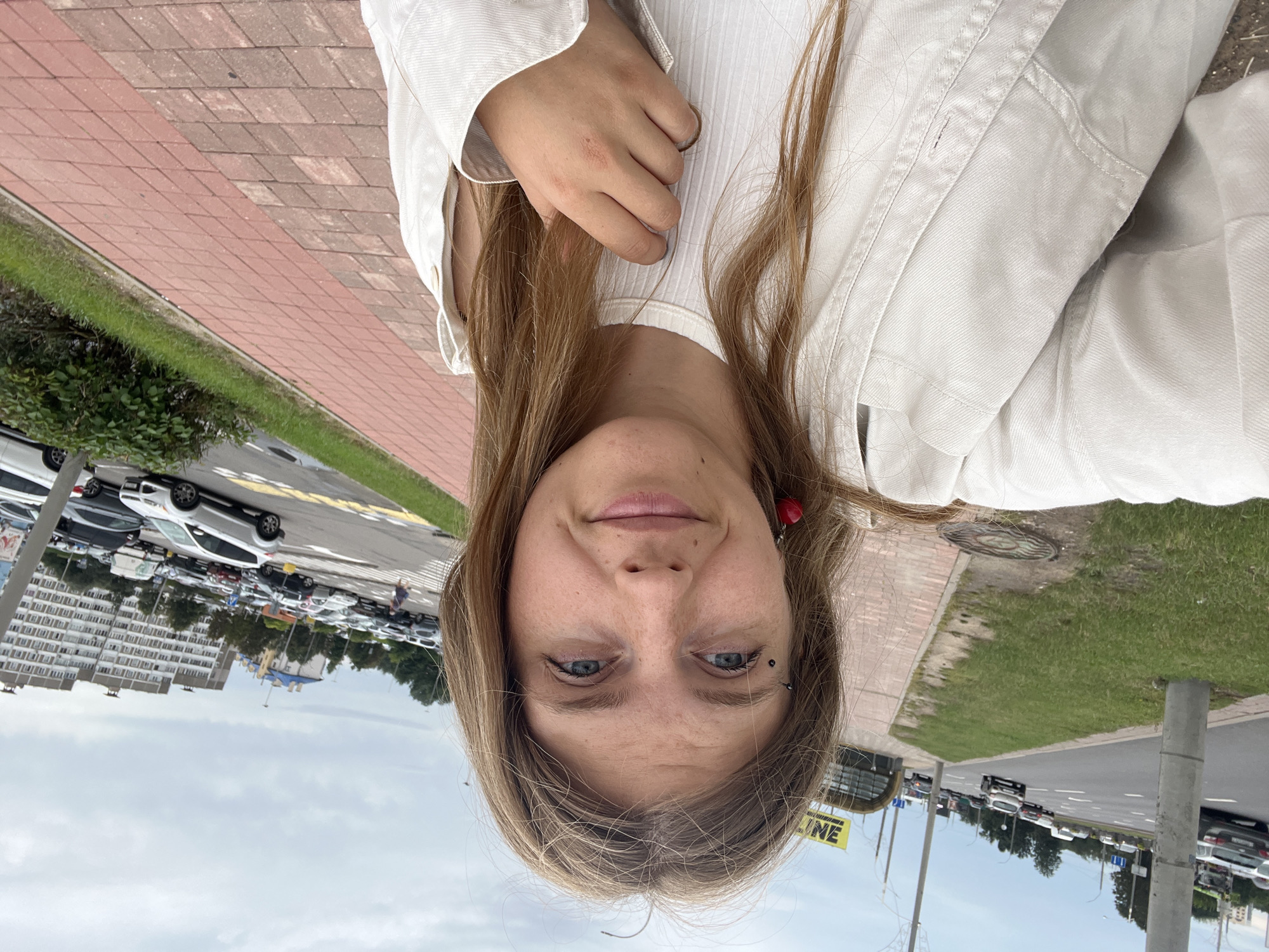 This is a selfie taken outdoors in Minsk, Belarus. The subject is a young woman with blonde hair, wearing a white top and a white jacket. She has a piercing above her left eye and is wearing a red earring. She is standing on a sidewalk with red brick paving, next to a grassy area and a road with cars. In the background, there is a parking lot filled with cars and several multi-story buildings. The sky is cloudy. A yellow sign with the word "LINE" is visible in the distance. The image appears to be taken during the daytime.