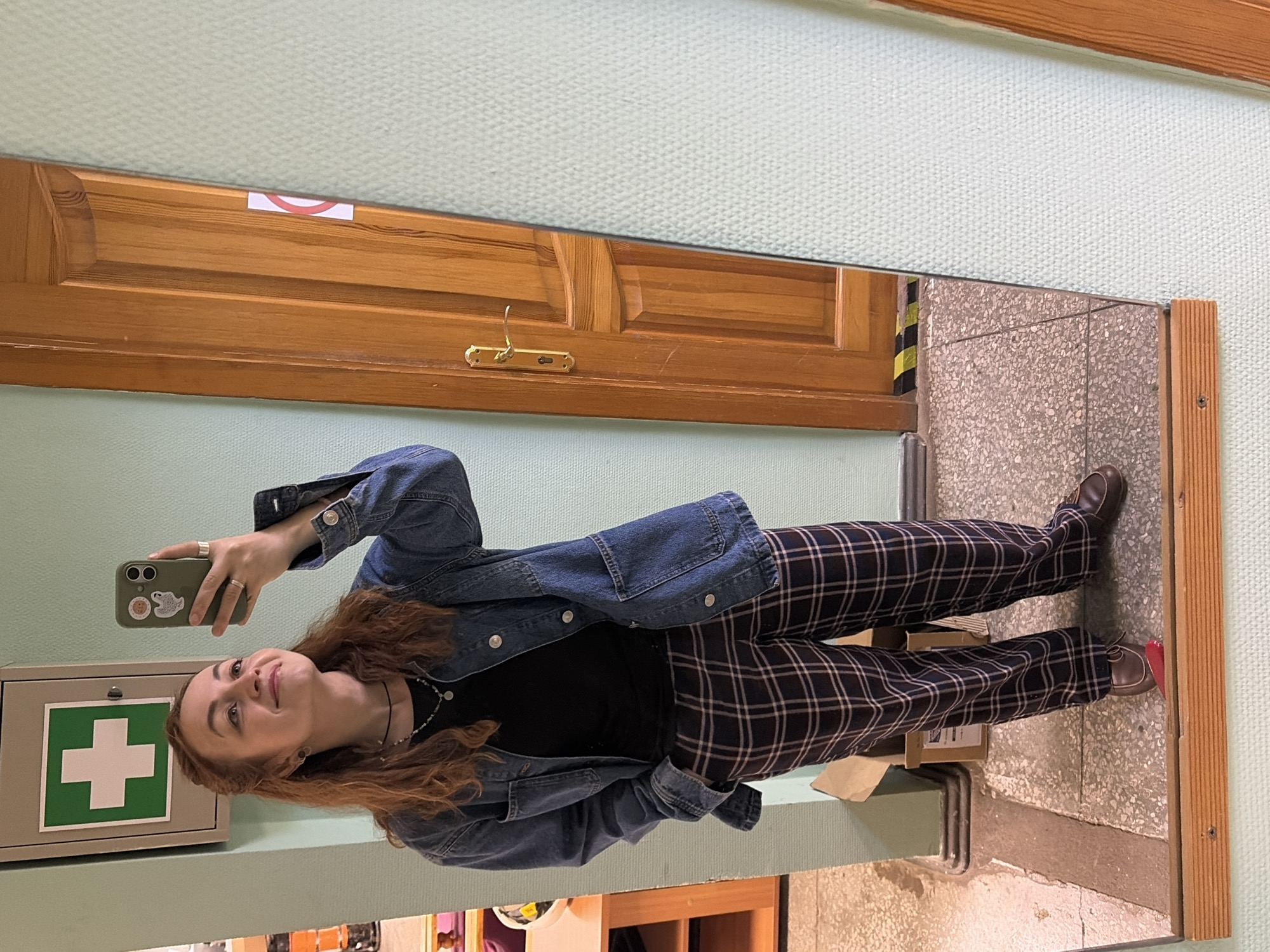 A young woman is taking a selfie in a mirror. She is wearing a black shirt, a denim jacket, and plaid pants. The background shows a wall with a green-painted surface. A wooden door is visible above her, as well as a first aid kit on the wall to her left. Based on the style of the first aid kit and architectural features, the location may be a public space in Chaikovsky, Russia. The reflection shows a tiled floor and a glimpse of an adjacent room.