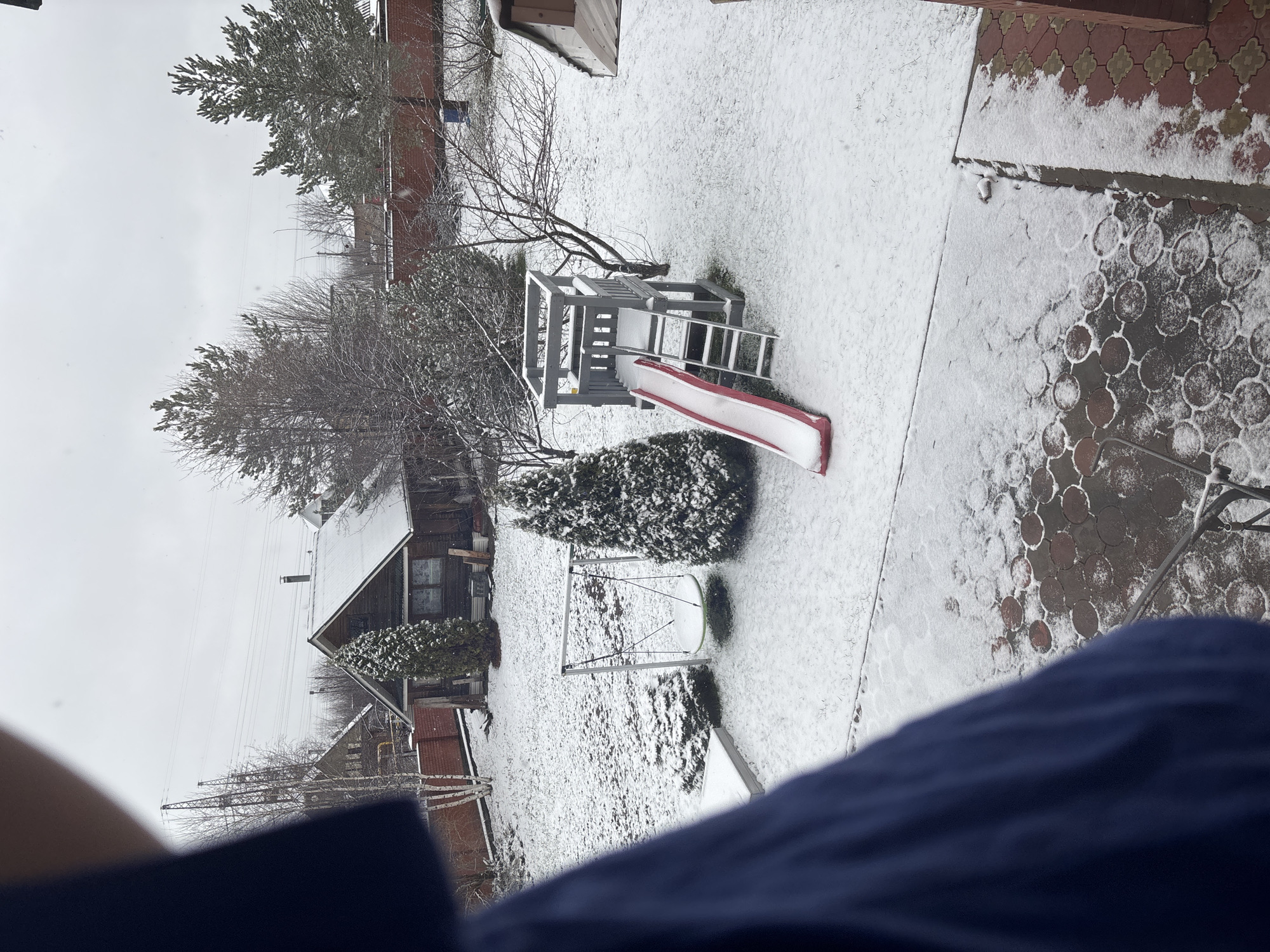 The image depicts a snow-covered backyard in Ферма, Russia. A wooden house with a gable roof is visible, along with a children's play structure that includes a slide. Other objects include a swing set, a snow-covered yard, brick walkways, and trees. The sky is overcast. A corner of a blue garment is visible in the foreground.