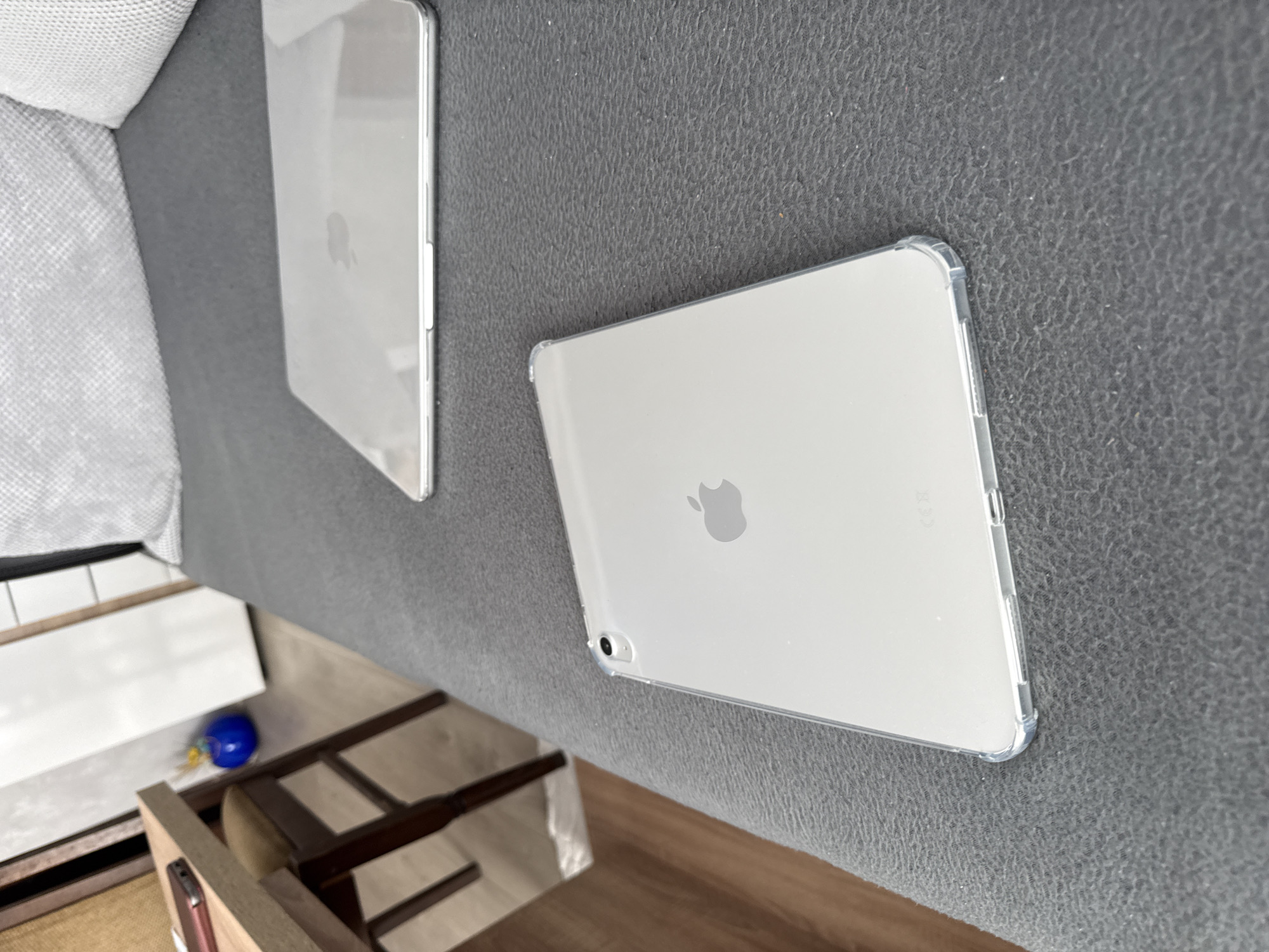 The image shows two silver Apple iPads lying on a gray fabric surface. One iPad is slightly out of focus in the background, while the other is in sharper focus in the foreground and appears to be in a clear plastic case. The Apple logos are visible on both devices. The lower left corner of the image includes a partially visible wooden structure. The image does not provide any information about the location in the city of Lovech, Bulgaria or any other location. There are no people present.