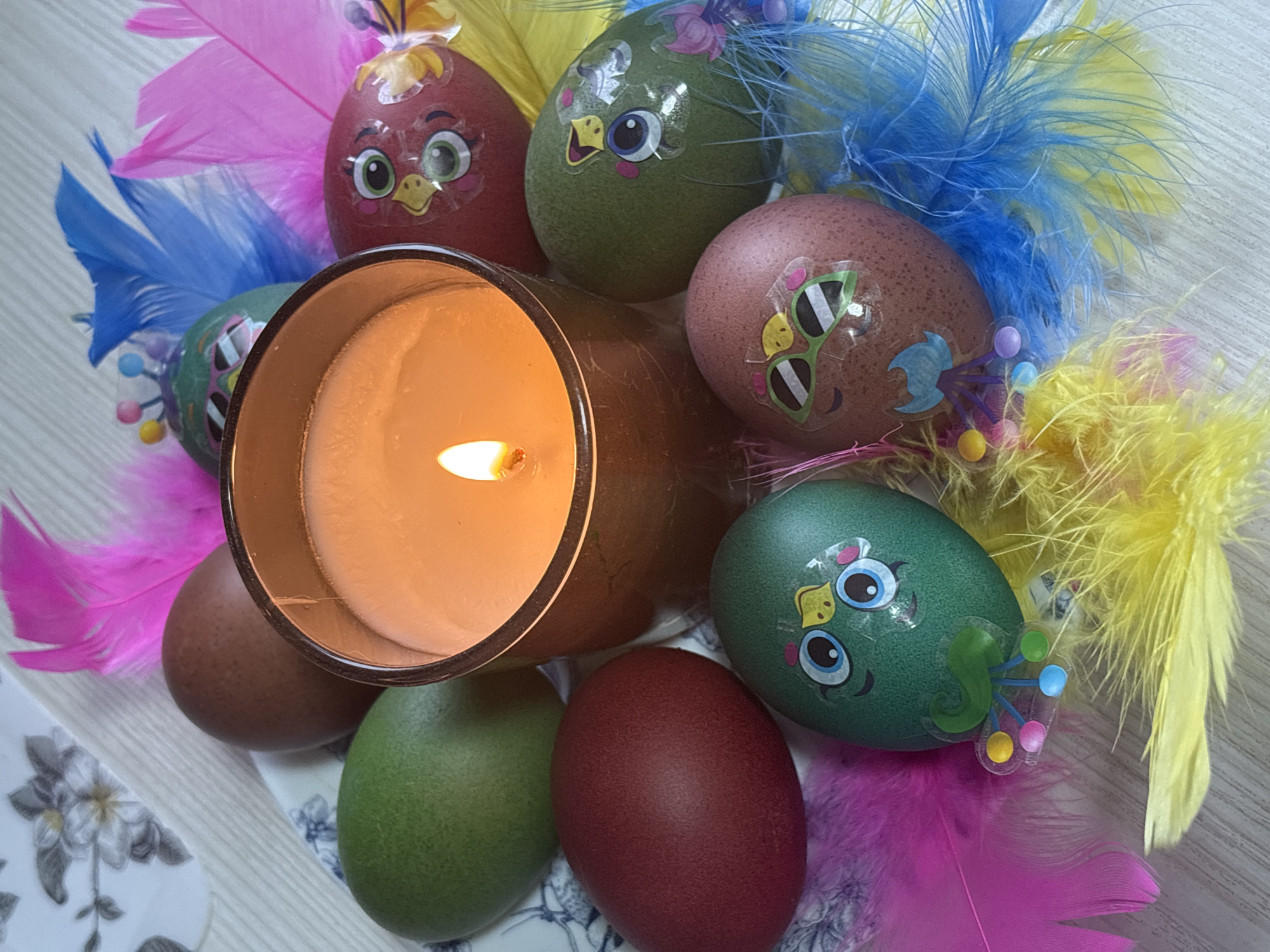 The image shows a lit candle in a gold-colored glass holder surrounded by decorated Easter eggs and colorful feathers. The eggs are various colors, including brown, green, and red, and feature cartoon-like stickers with bird-like faces and other decorative elements. The feathers are pink, blue, and yellow. The arrangement is placed on a surface with a floral pattern. There is no indication of Chaikovsky, Russia.